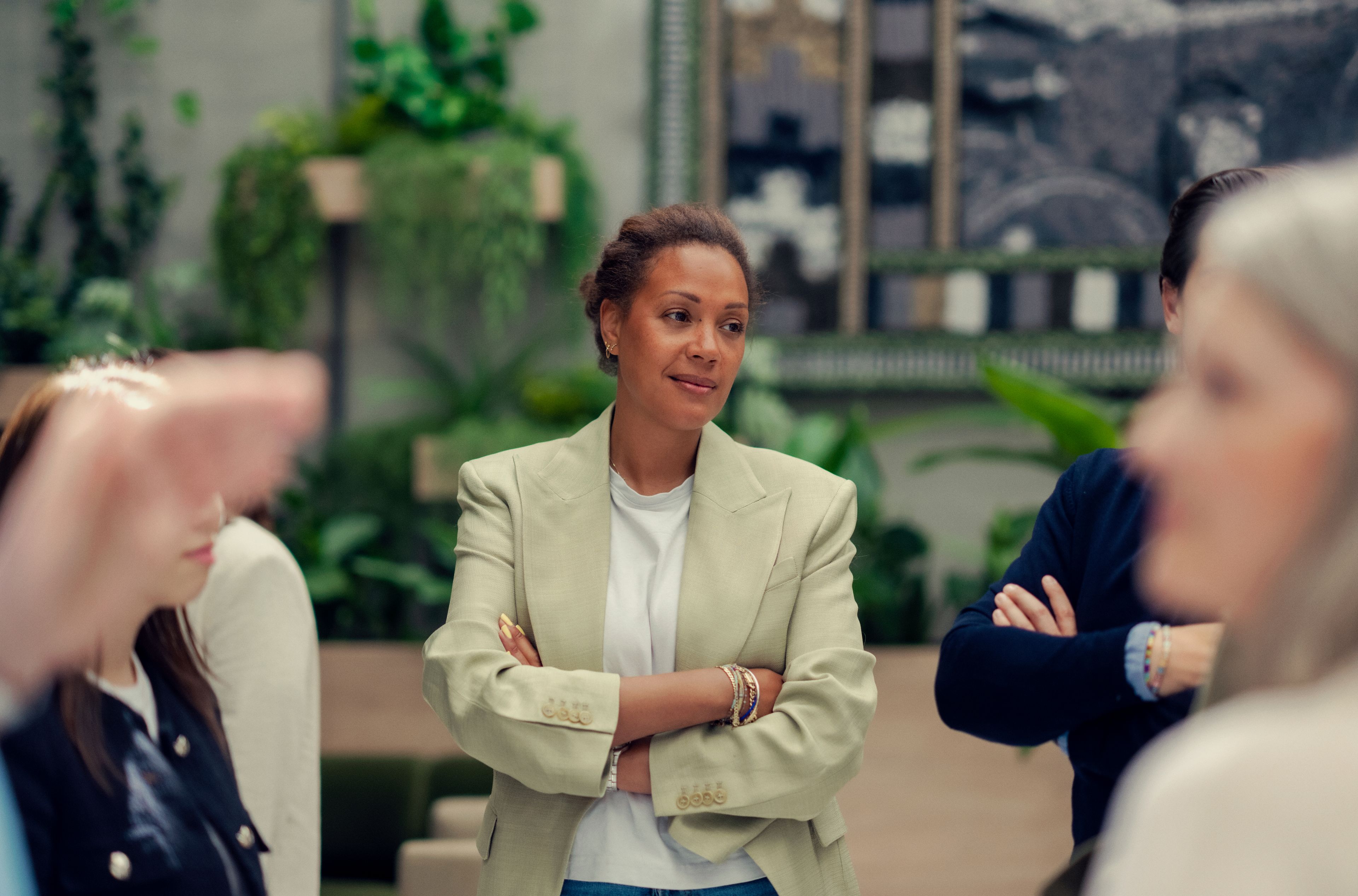 En kvinne i en lett blazer står med armene i kors og observerer oppmerksomt en diskusjon i et rom med planter og abstrakt kunst. Stemningen er tankefull.