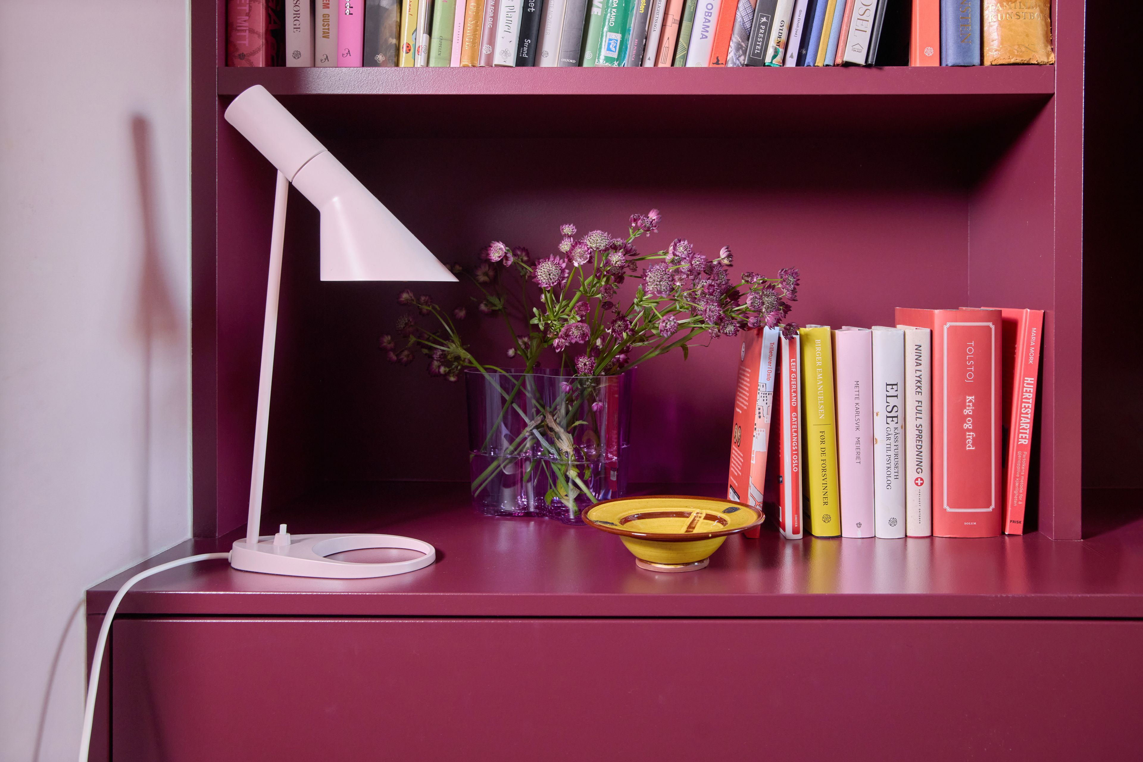Plommefarget bokhylle med hvit AJ-bordlampe, lilla vase med markblomster og bøker i rosa og oransje toner.