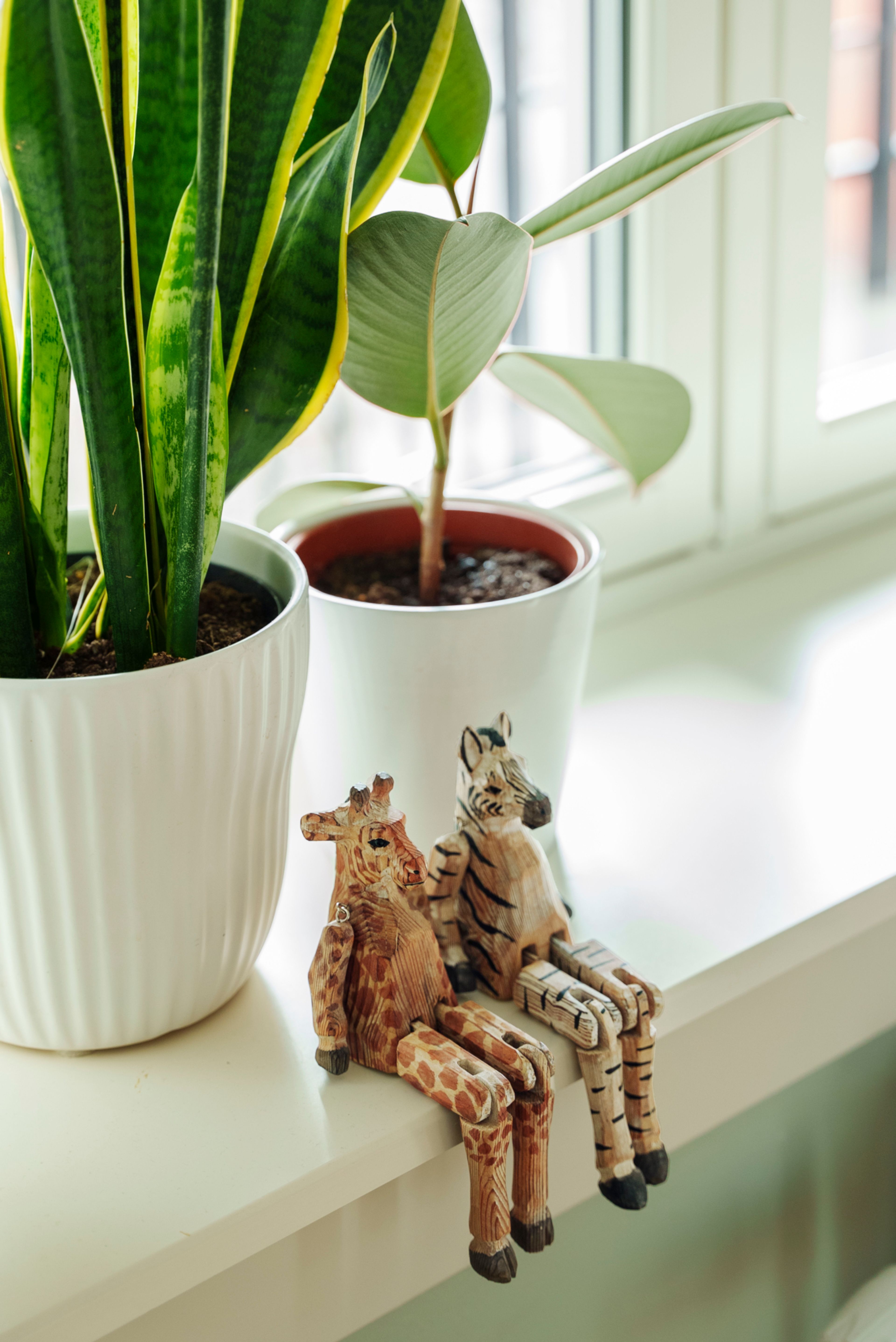 To små dekorsjiraffer sittende i en vinduskarm, med to planter bak seg.