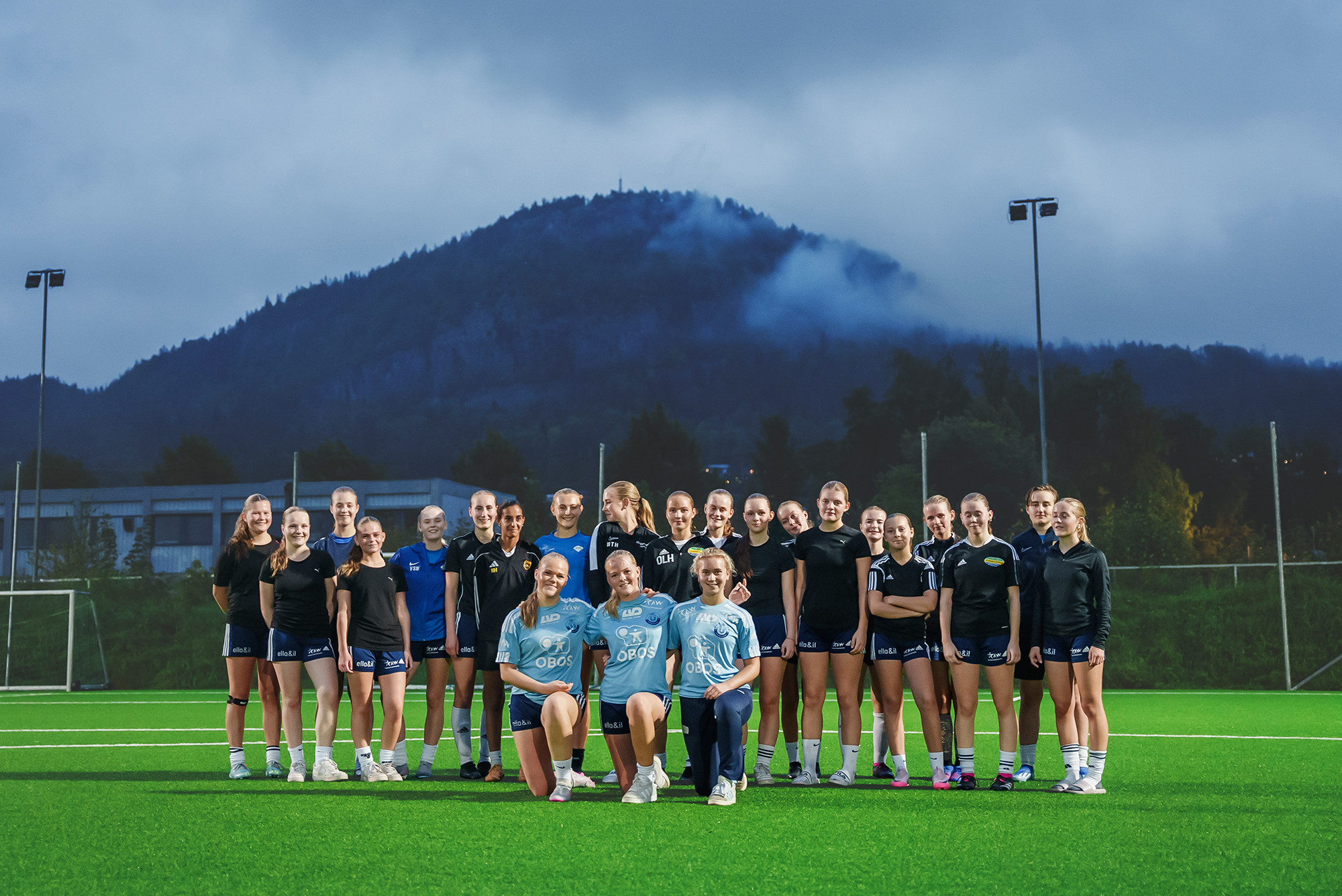 Lagbilde av jenter-17-gruppen i Union Bærum Kvinner. De er ute på en kunstgressbane mot en grå himmel og fjell i bakgrunnen.