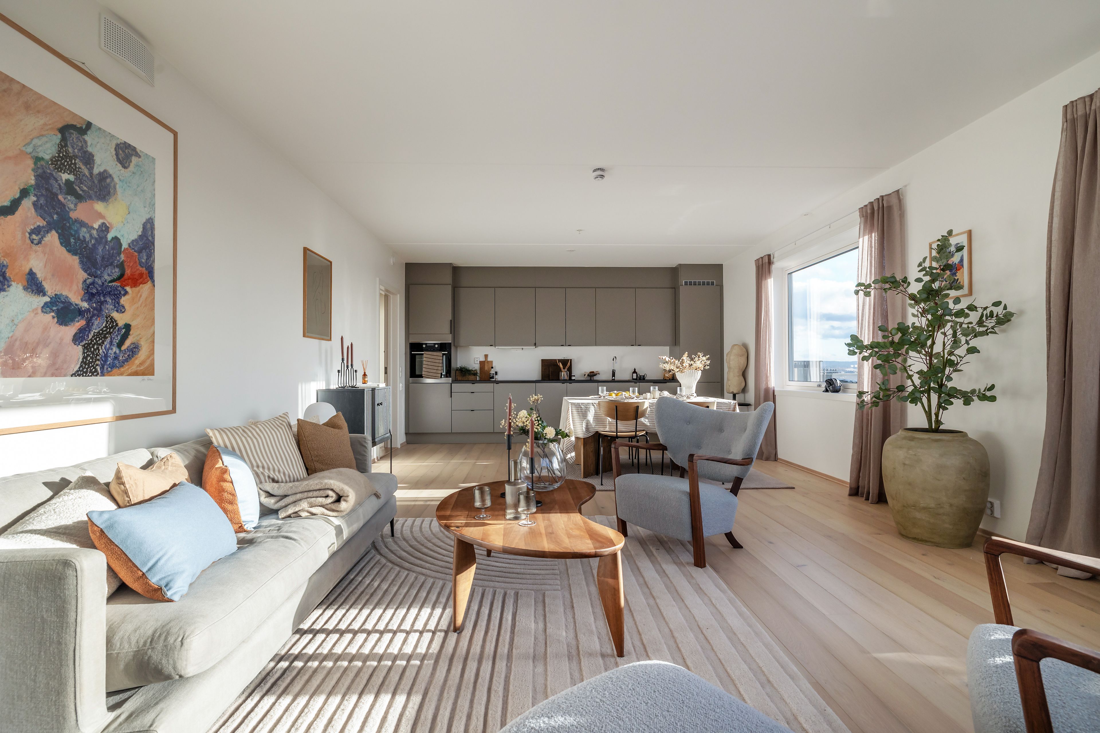 Åpen løsning med stue og kjøkken i Løren botaniske, innredet i naturfarger og lyseblått. I forgrunnen er det stue med sofa, trebord og stoler, videre en spiseplass og innerst en kjøkkeninnredning på kortsiden av rommet.