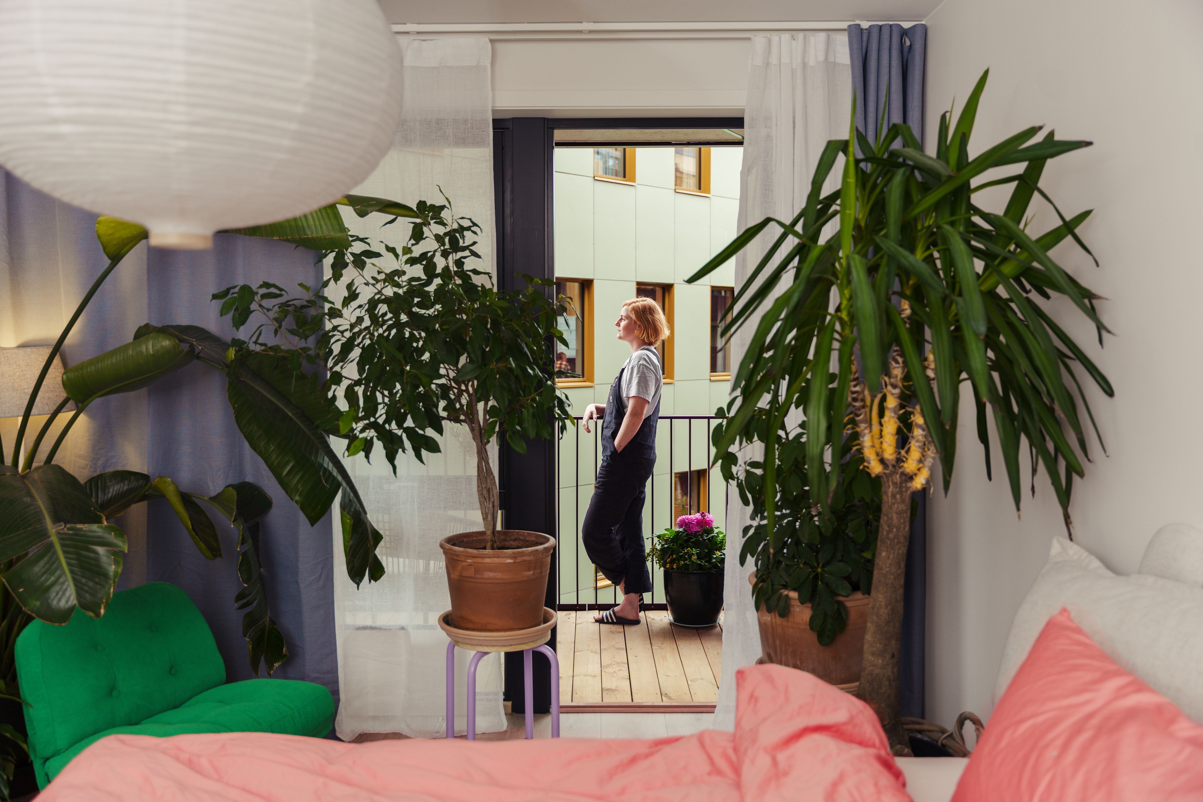 Kvinne står på verandaen, mens vi ser gjennom rommet hennes, som har store planter og fargerike møbler