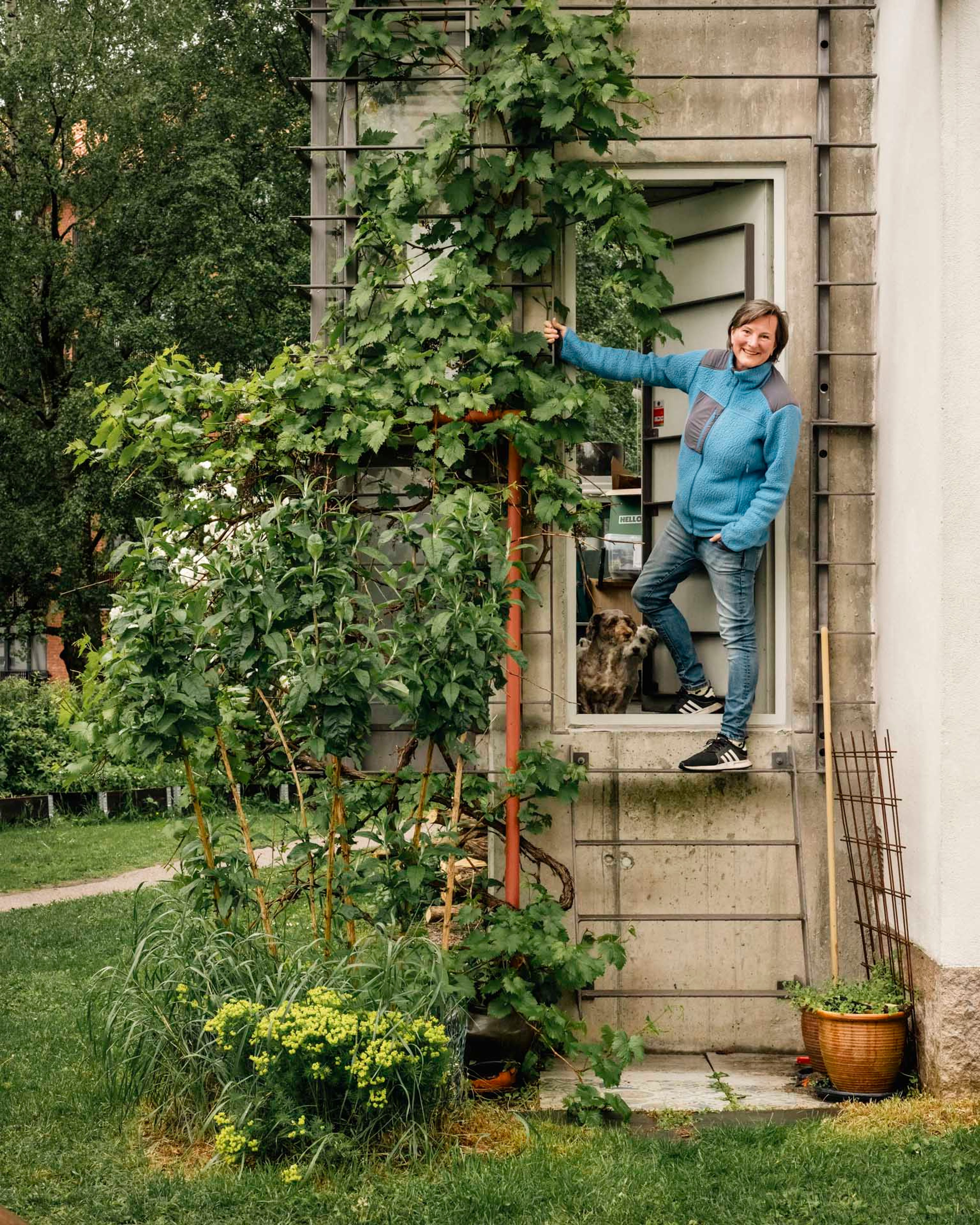 Smilende kvinne i blå genser holder seg i en vinstokk ved inngangsdøren til boligen sin, med en hund synlig innenfor og en frodig hage rundt.