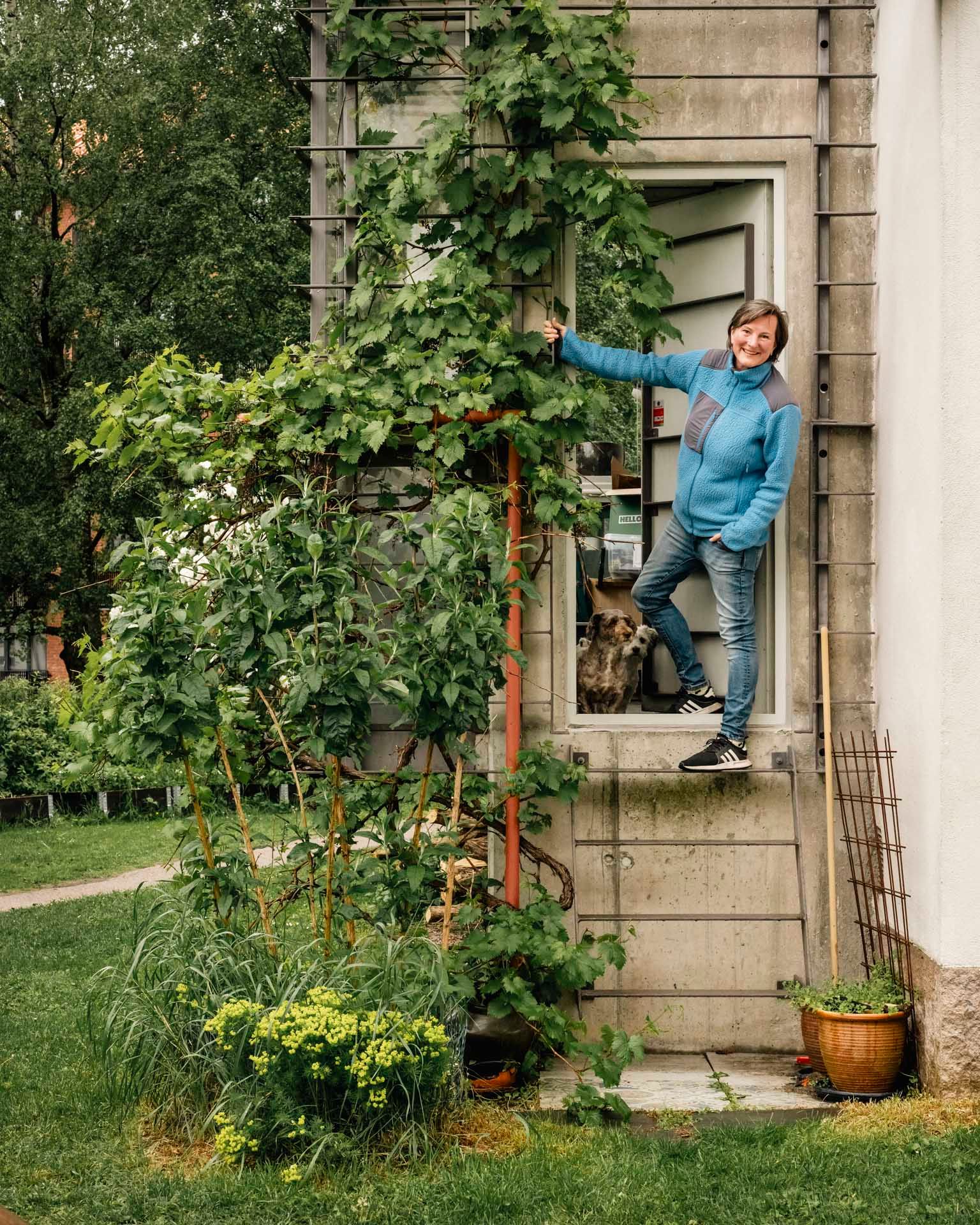 Smilende kvinne i blå genser holder seg i en vinstokk ved inngangsdøren til boligen sin, med en hund synlig innenfor og en frodig hage rundt.