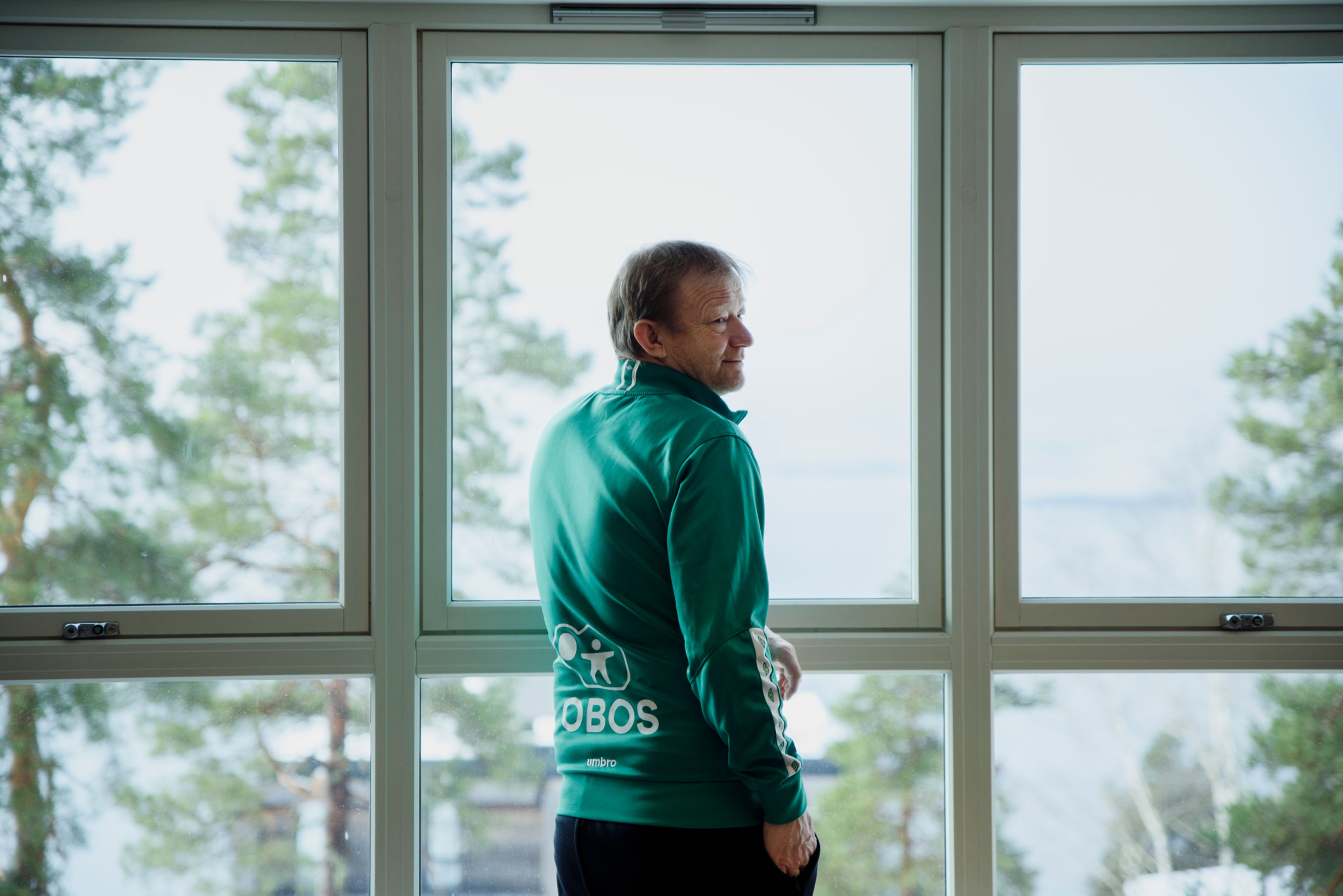 Bilde av Geir på innsiden av Sunnaas sykehus. Han lener seg mot vinduet og har på en grønn treningsjakke med logen til OBOS.