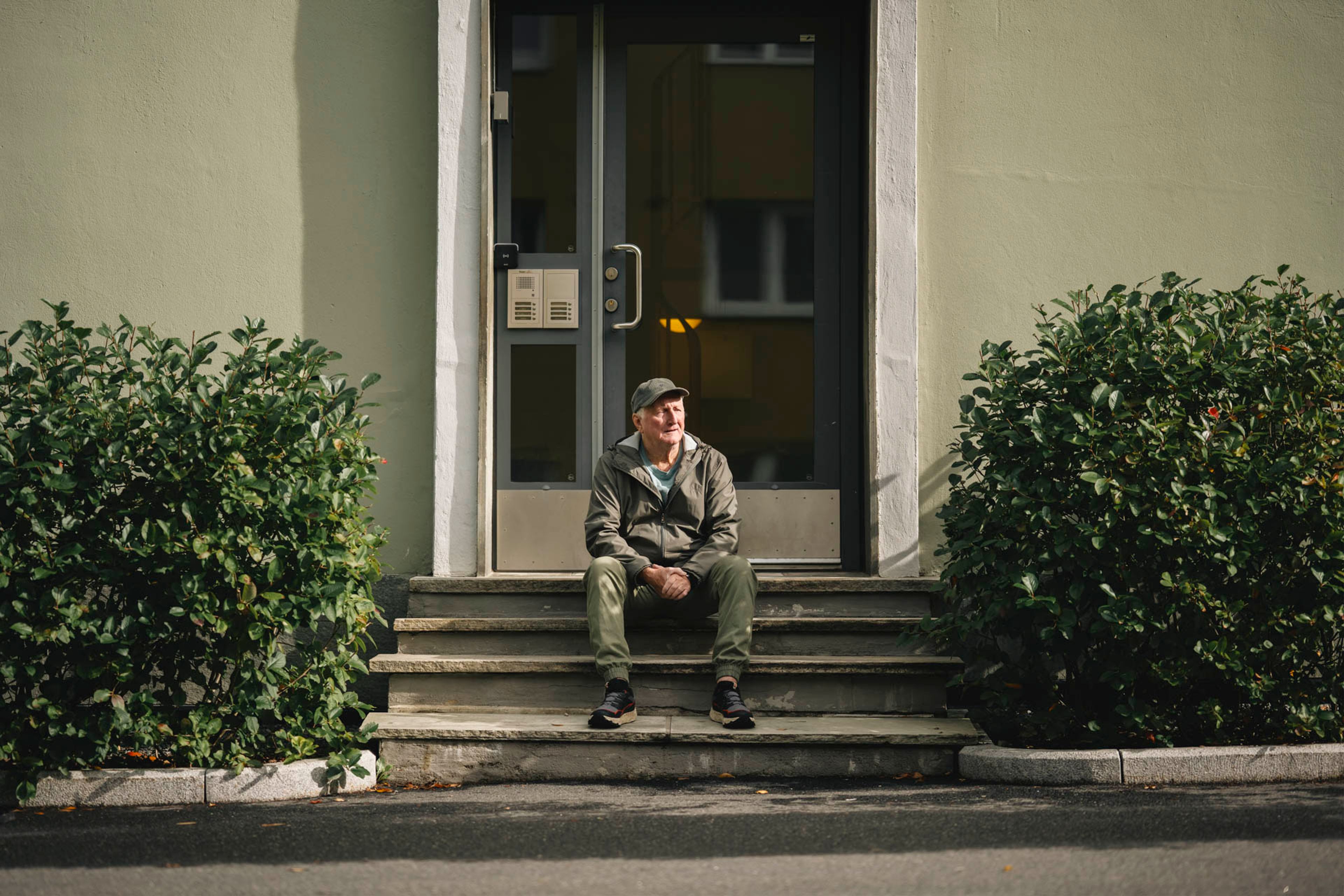 En eldre mann sitter på trappen foran inngangsdøren til en bygård, omgitt av grønne busker.
