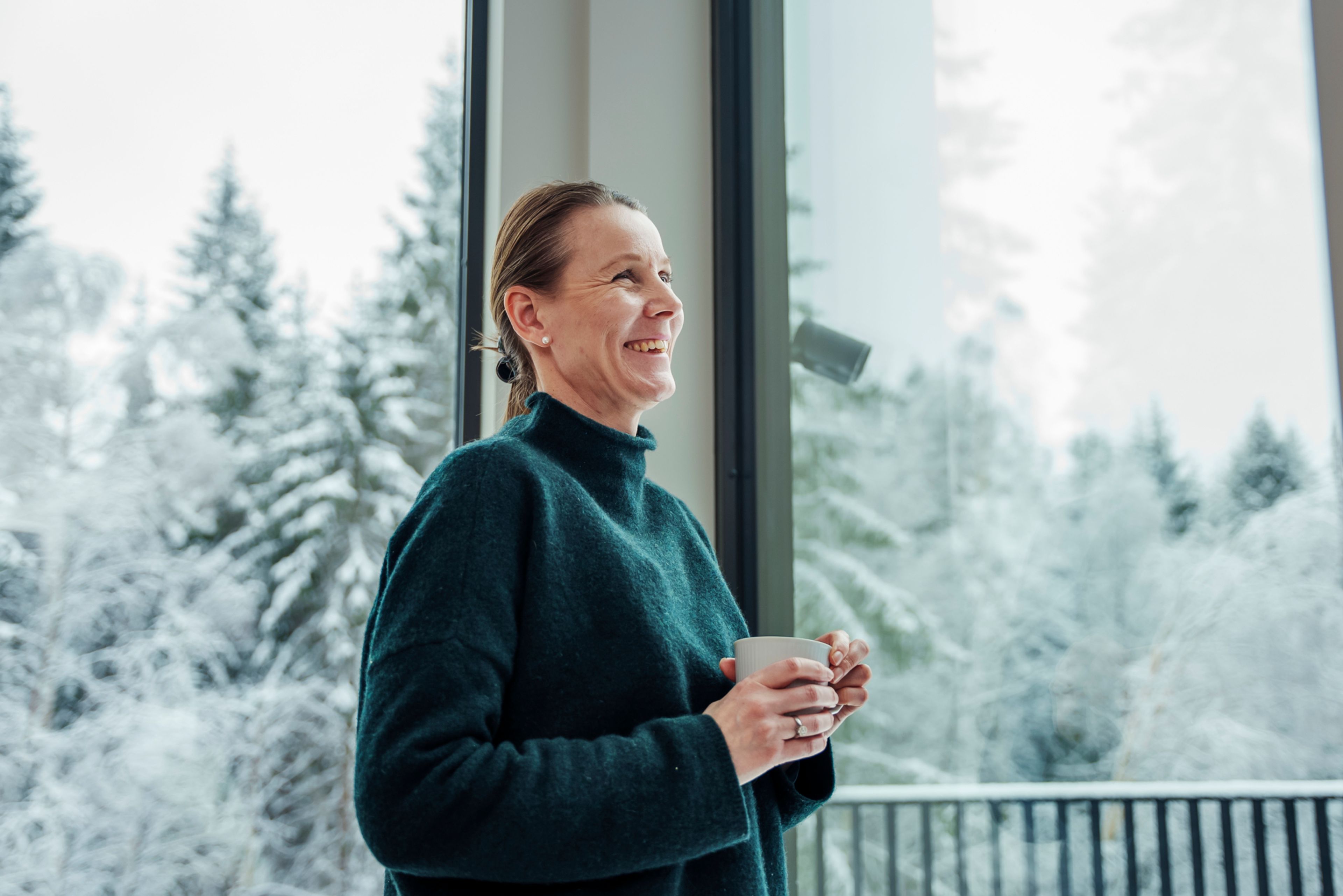 Nina står og ser ut av vinduet mens hun holder en kaffekopp.