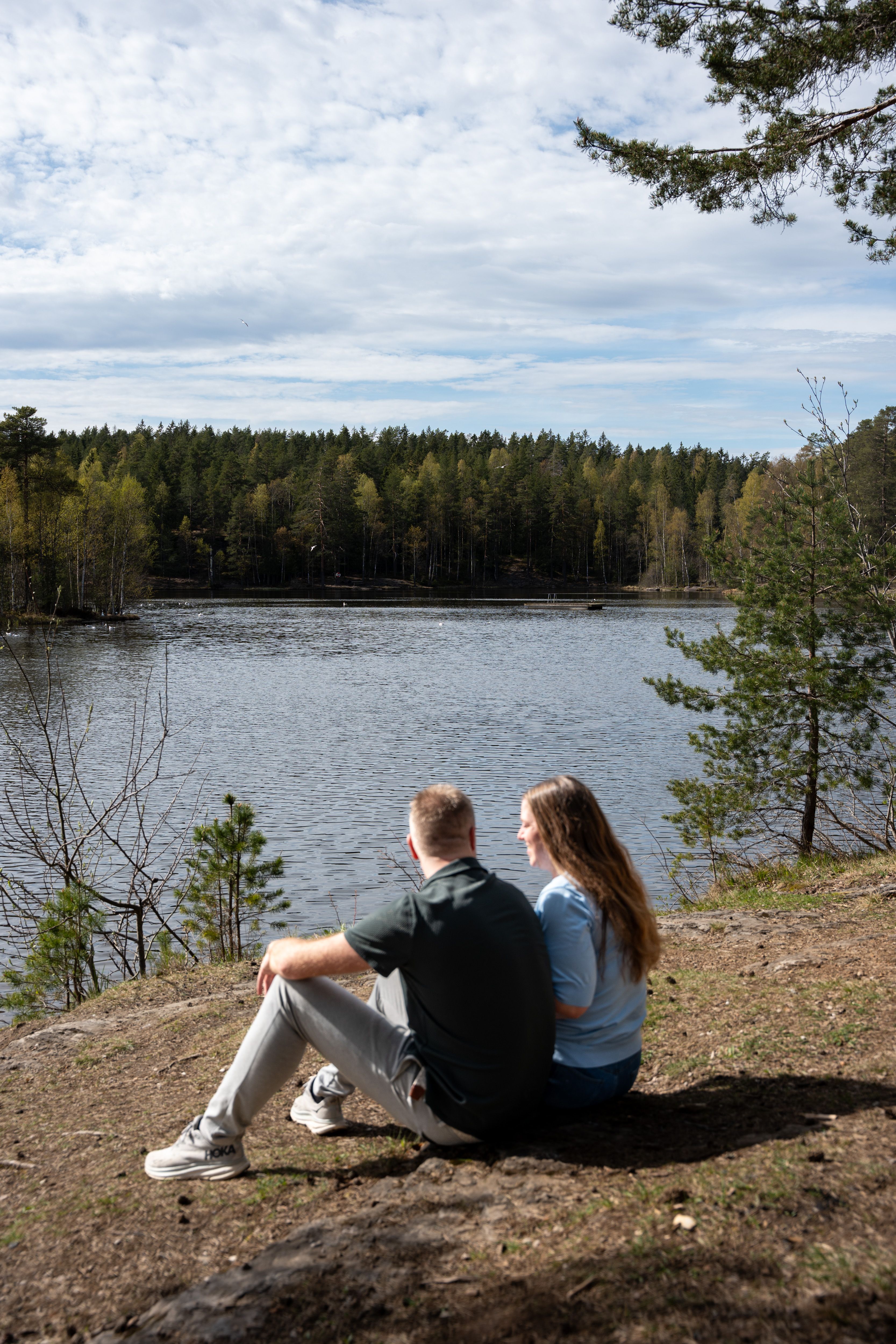 Et par sitter på en bergknaus ved bredden av et rolig vann omgitt av skog.
