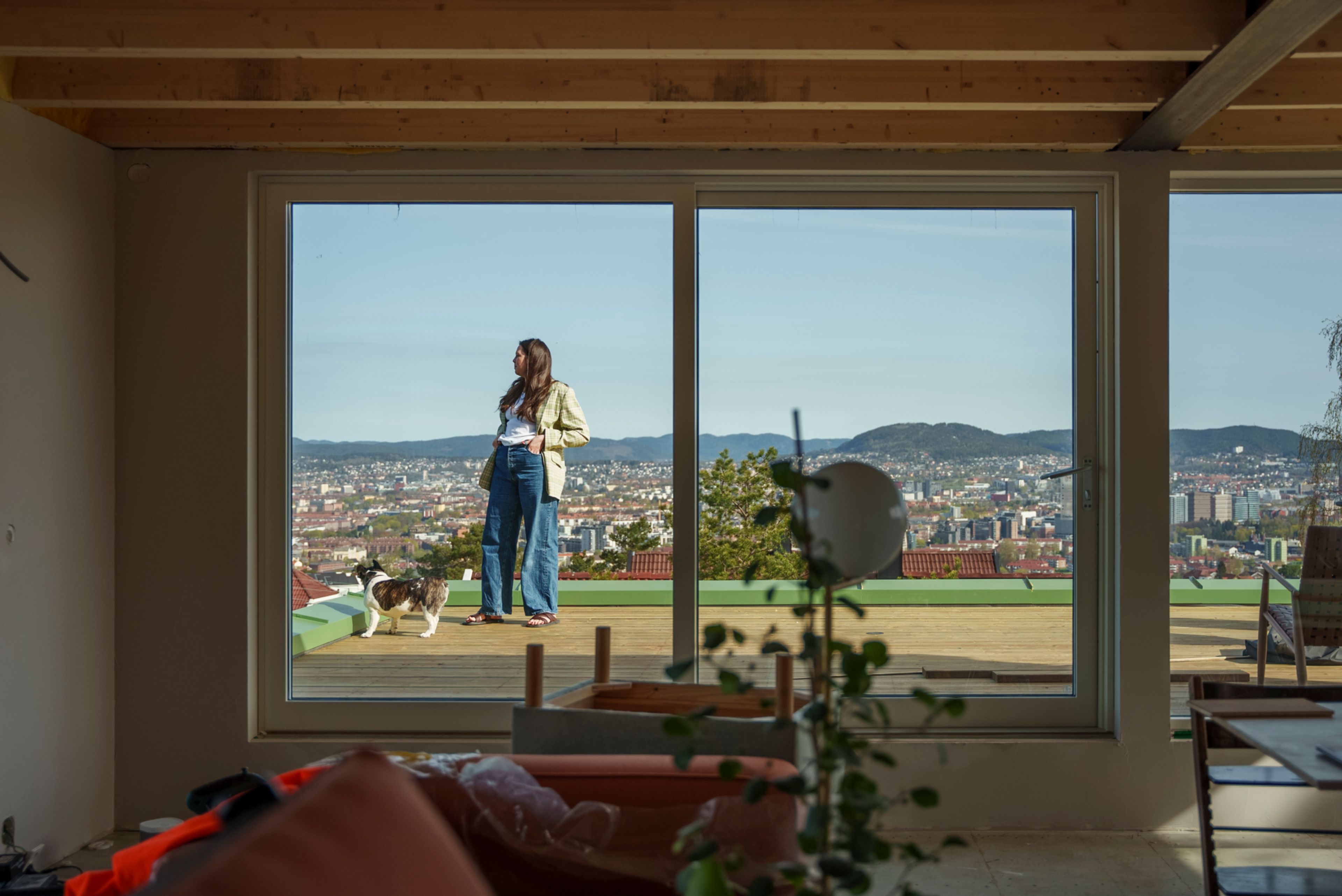 Bilde av en kvinne som står på en veranda. Bildet er tatt fra gjennom vinduet på innsiden av huset. Vi ser utsikt over Oslo i bakgrunnen.