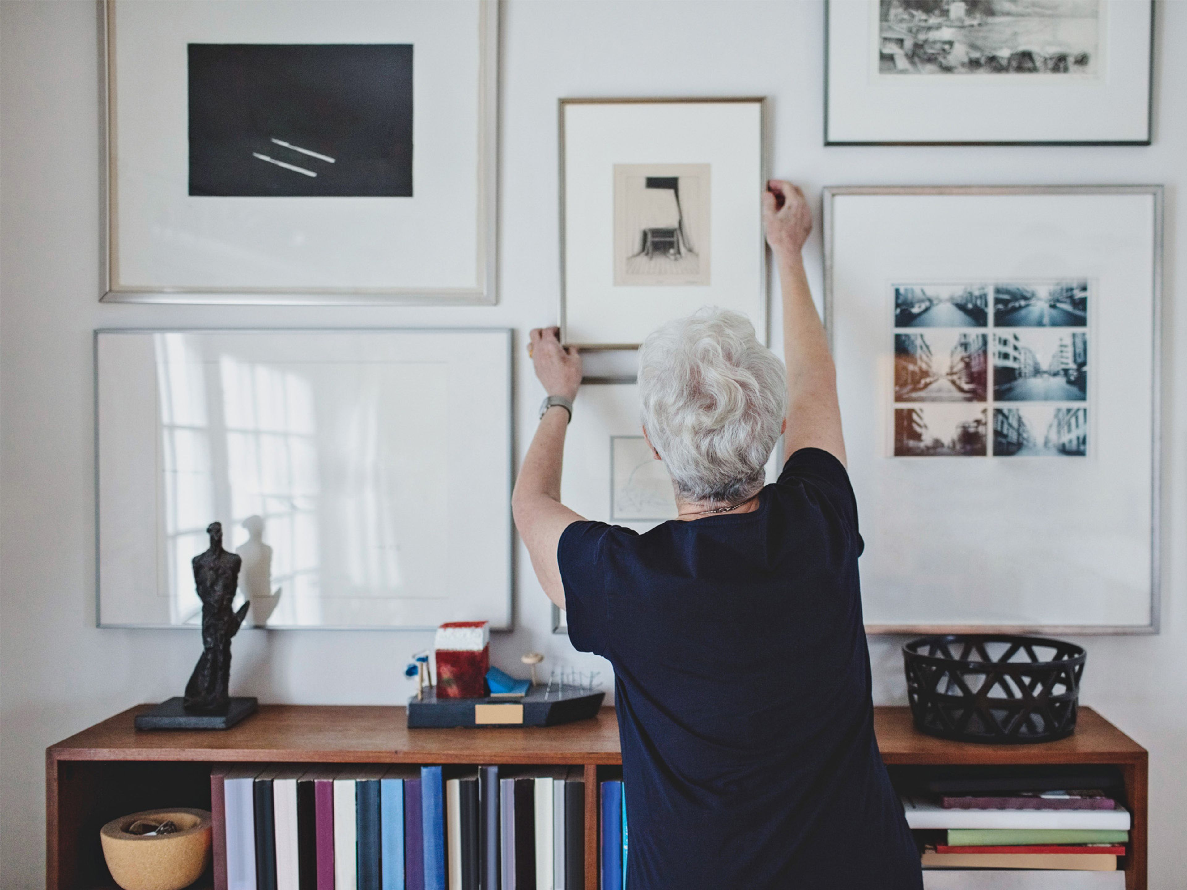 En &auml;ldre dam h&auml;nger upp tavlor p&aring; en tavelv&auml;gg
