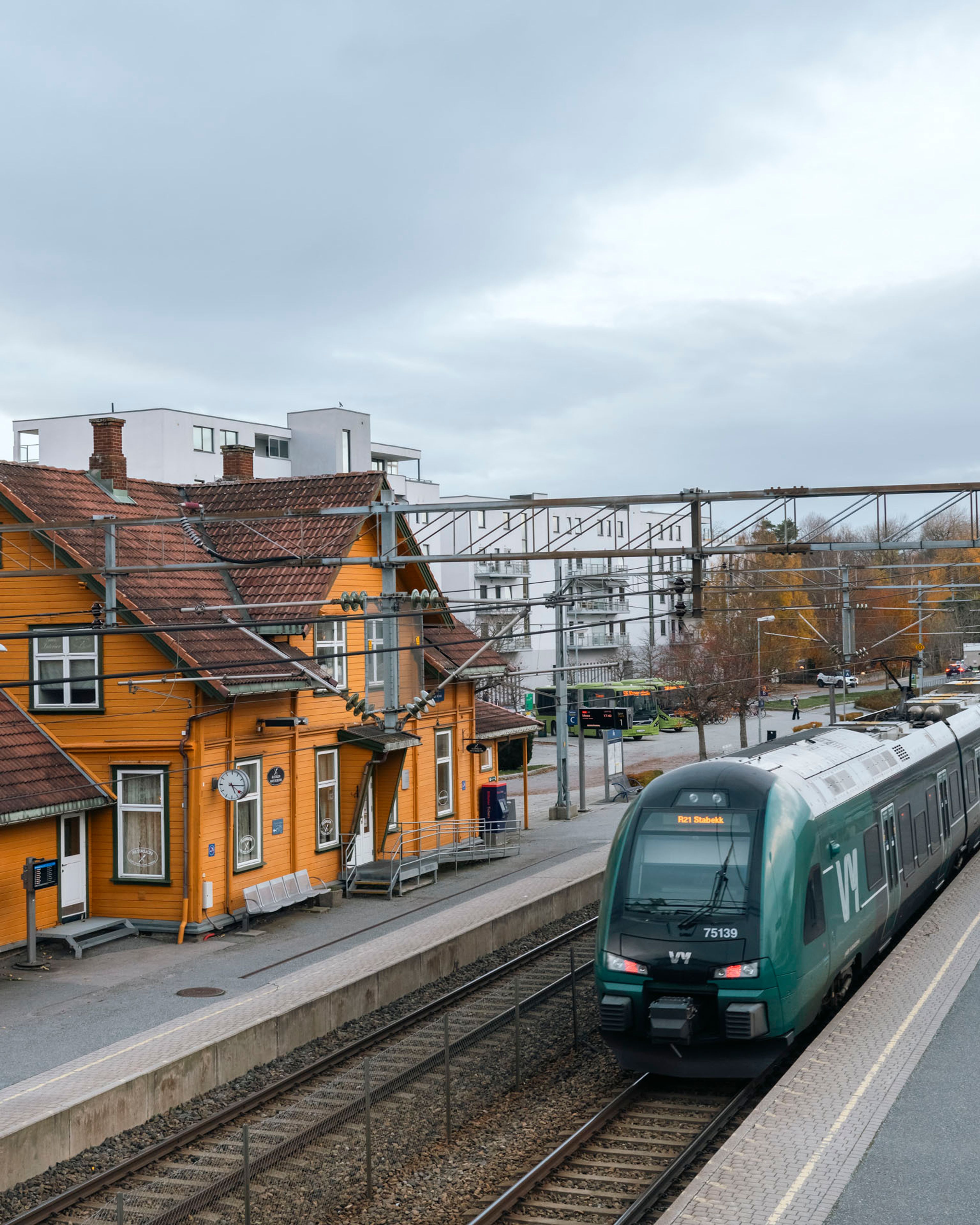 Foto av Ås stasjon, med oransje stasjonsbygg i tre, og et tog stående på et av sporene.