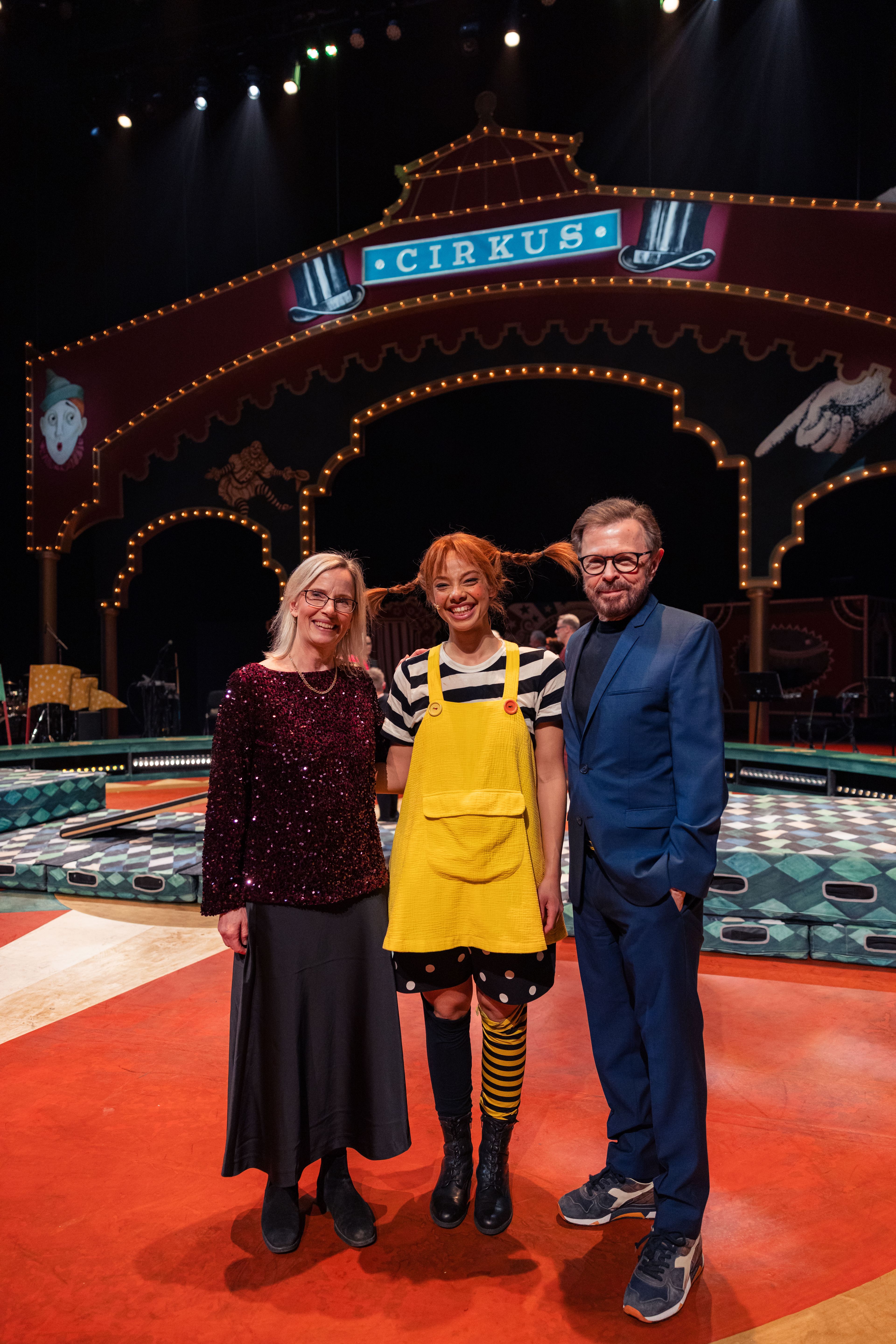 Ulvaeus og skuespilleren i Pippi-kostyme står sammen med en kollega foran et opplyst sirkustelt av tre på scenen.