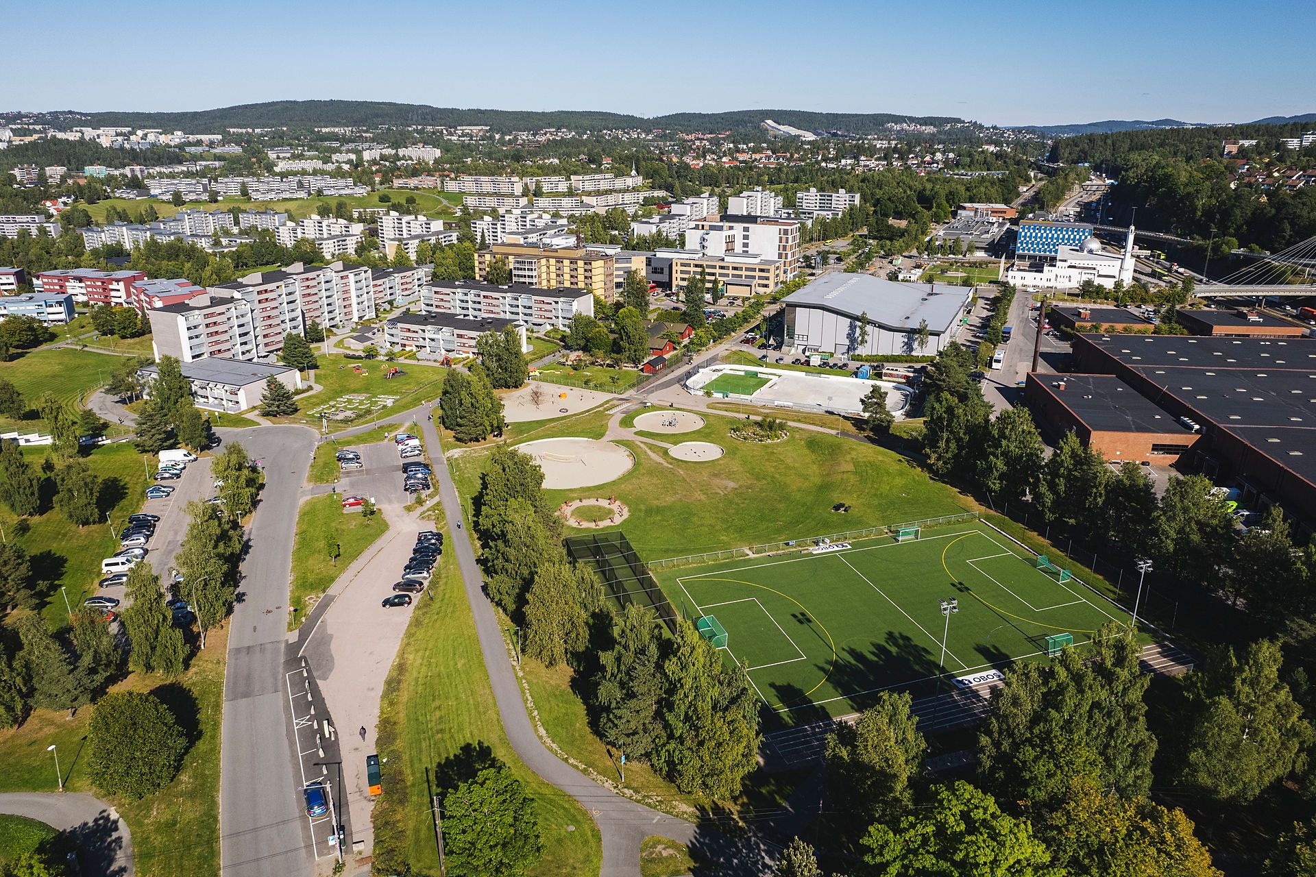 Et dronebilde av hele bydelen, hvor man ser en stor fortballbane, nærheten til marka og flere boligblokker.