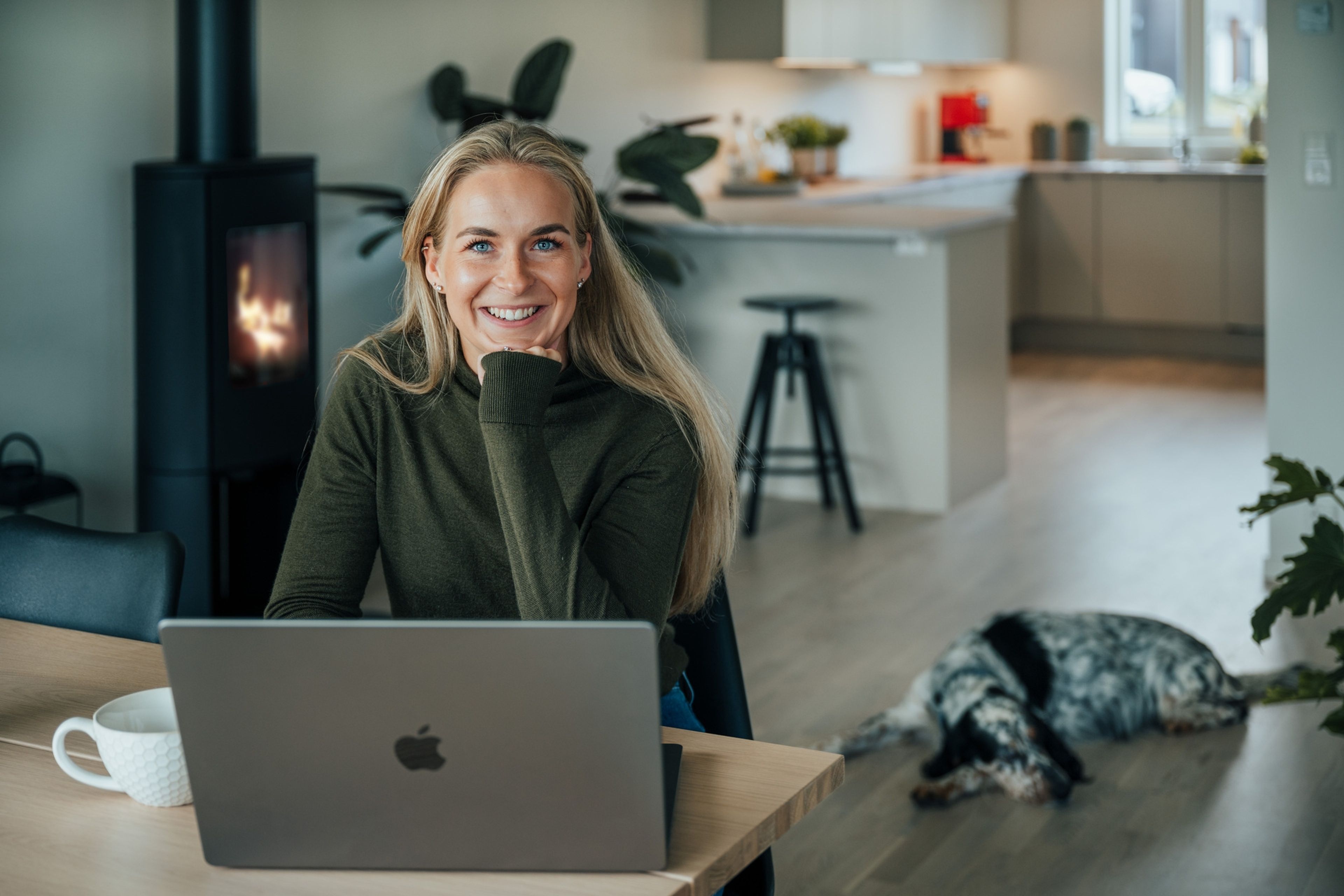 En ung kvinne med langt lyst hår sitter foran en mac i leiligheten sin. I bakgrunnen ligger det en hund på gulvet.
