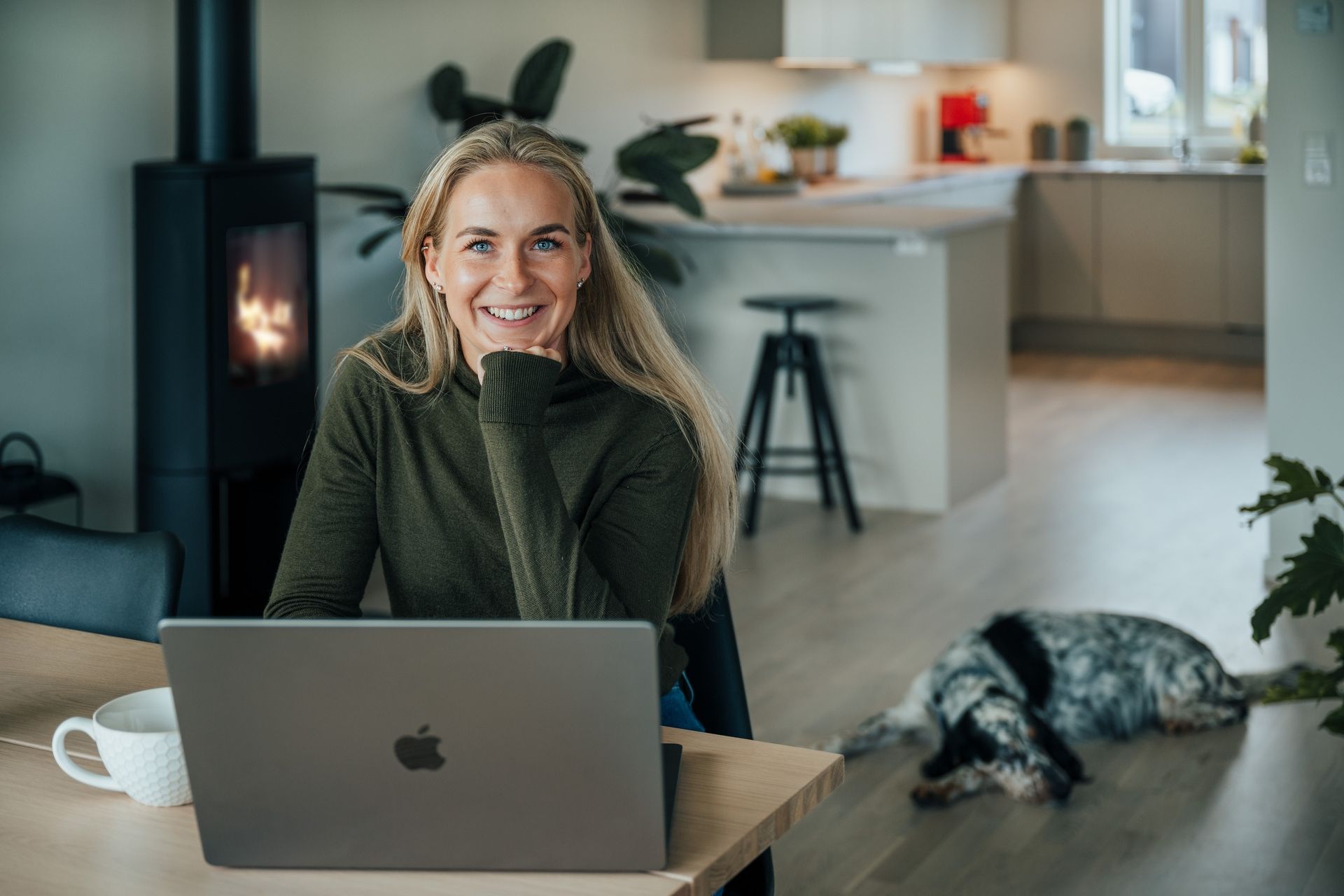 En ung kvinne med langt lyst hår sitter foran en mac i leiligheten sin. I bakgrunnen ligger det en hund på gulvet.