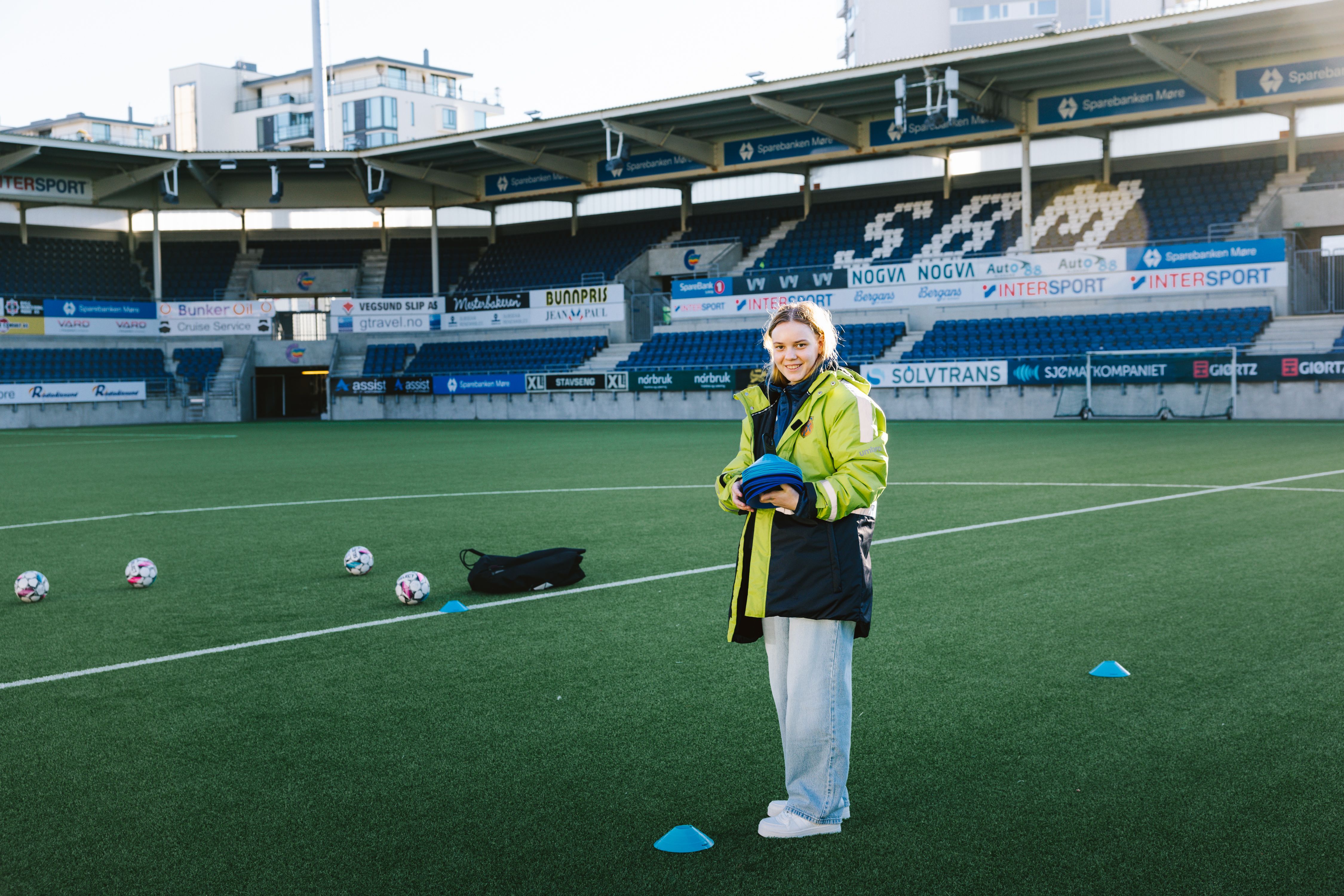 Kristina står midt på fotballbanen med en utstyrsbag og blå kjegler ved siden av, klar til å sette opp før kampstart.