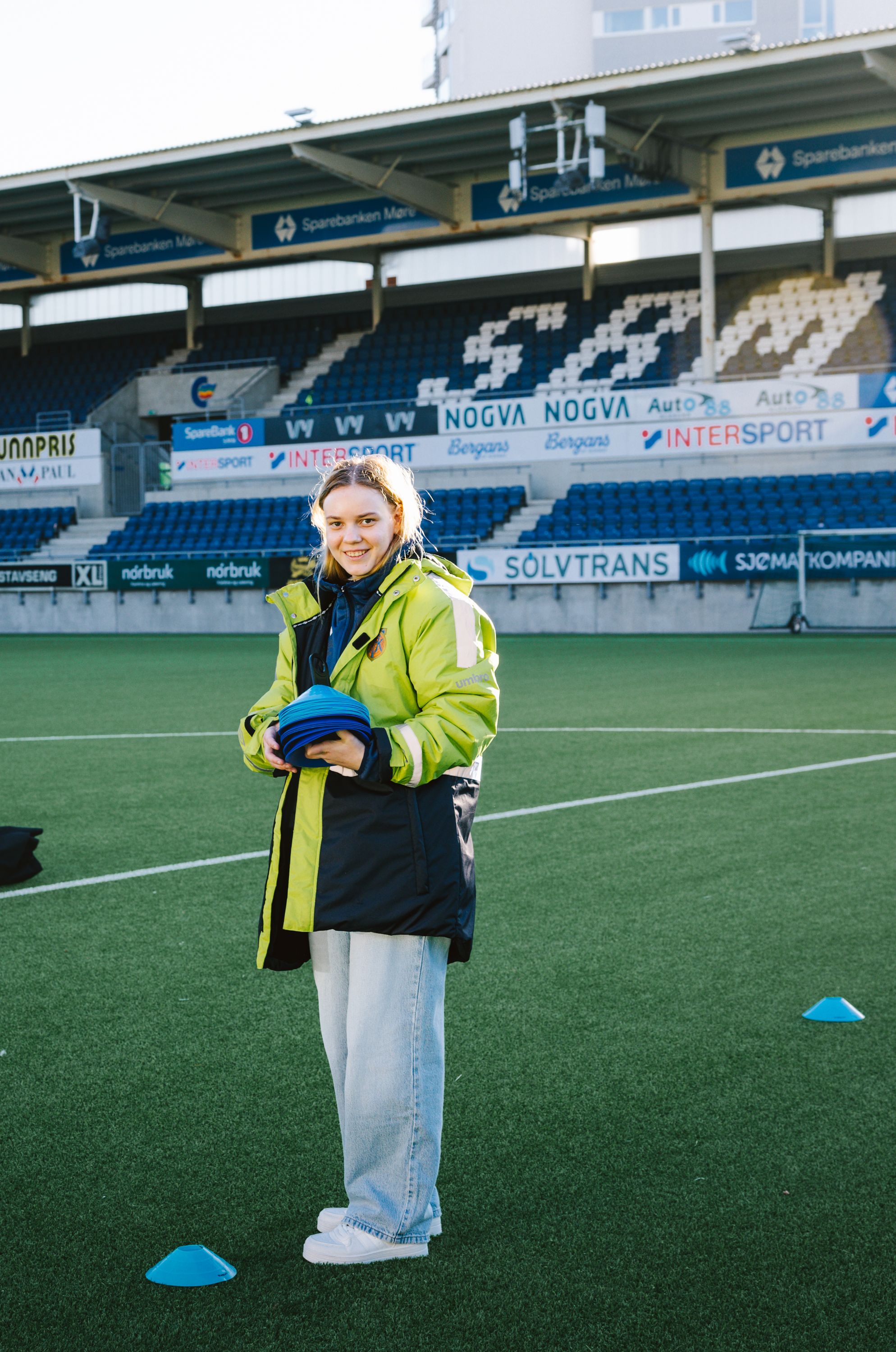 Kristina står midt på fotballbanen med en utstyrsbag og blå kjegler ved siden av, klar til å sette opp før kampstart.