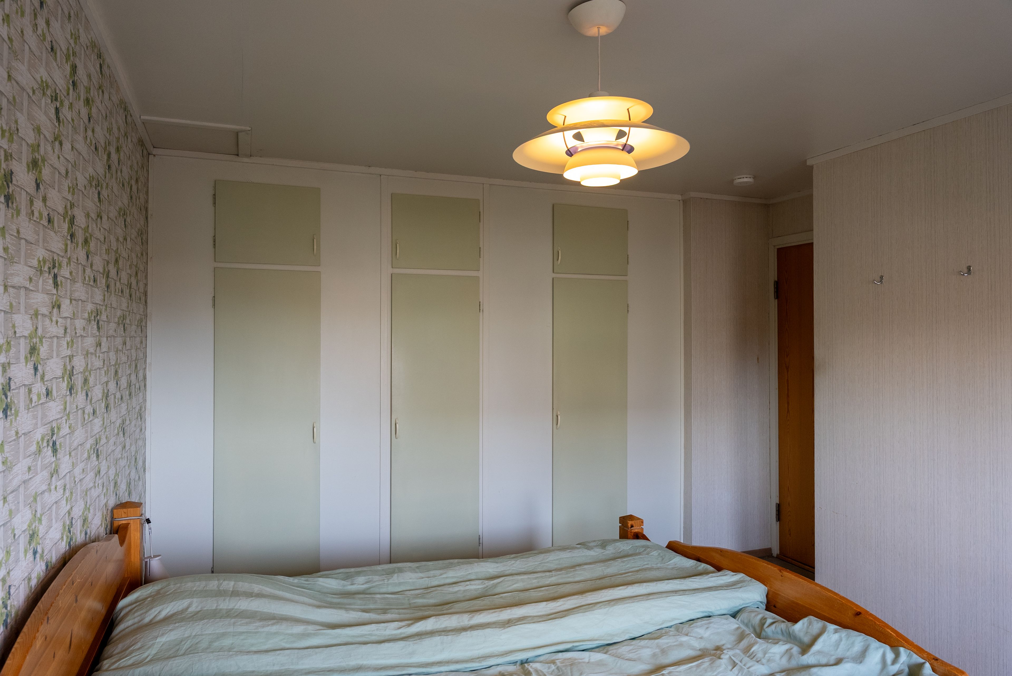 Eldre soverom med blomstrete tapet, linoleumsgulv, klesskap og furuseng.