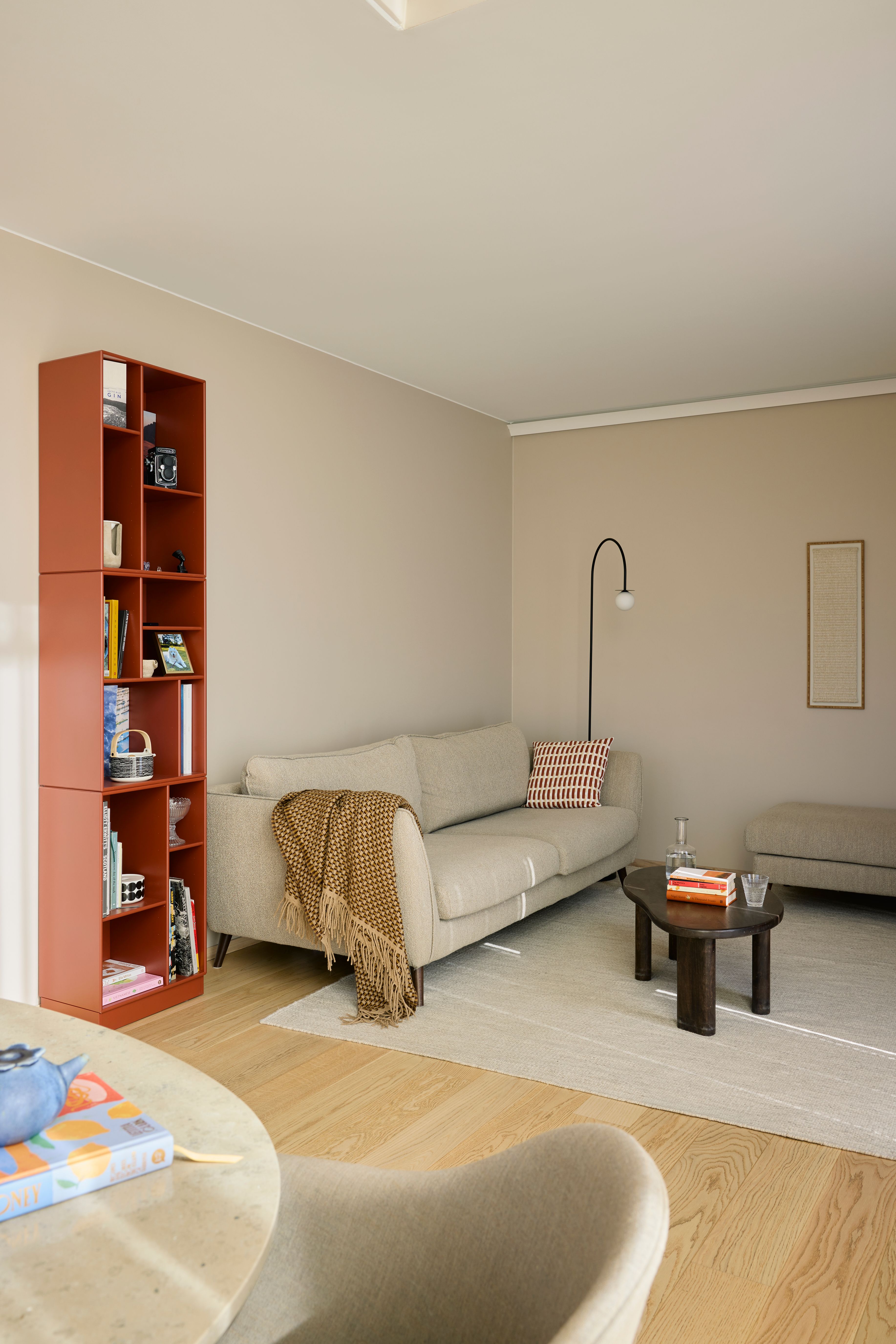 Stue med høy rustrød bokhylle, beige sofa med strikket pledd og stripete pute, buet gulvlampe og lite rundt sofabord i tre.