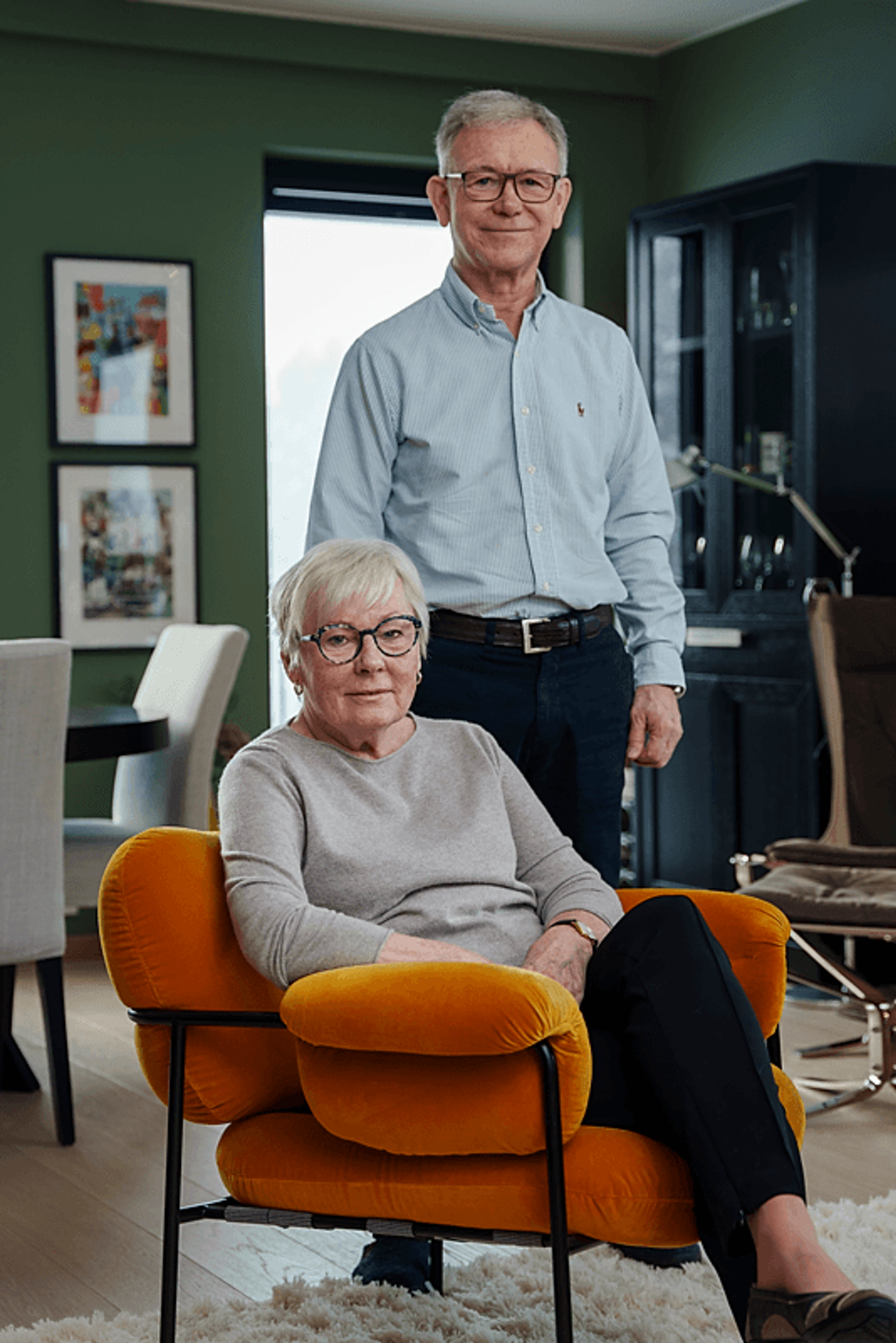 Elsa og Tore i stua si, Elsa sittende i en oransje stol med Tore stående bak henne