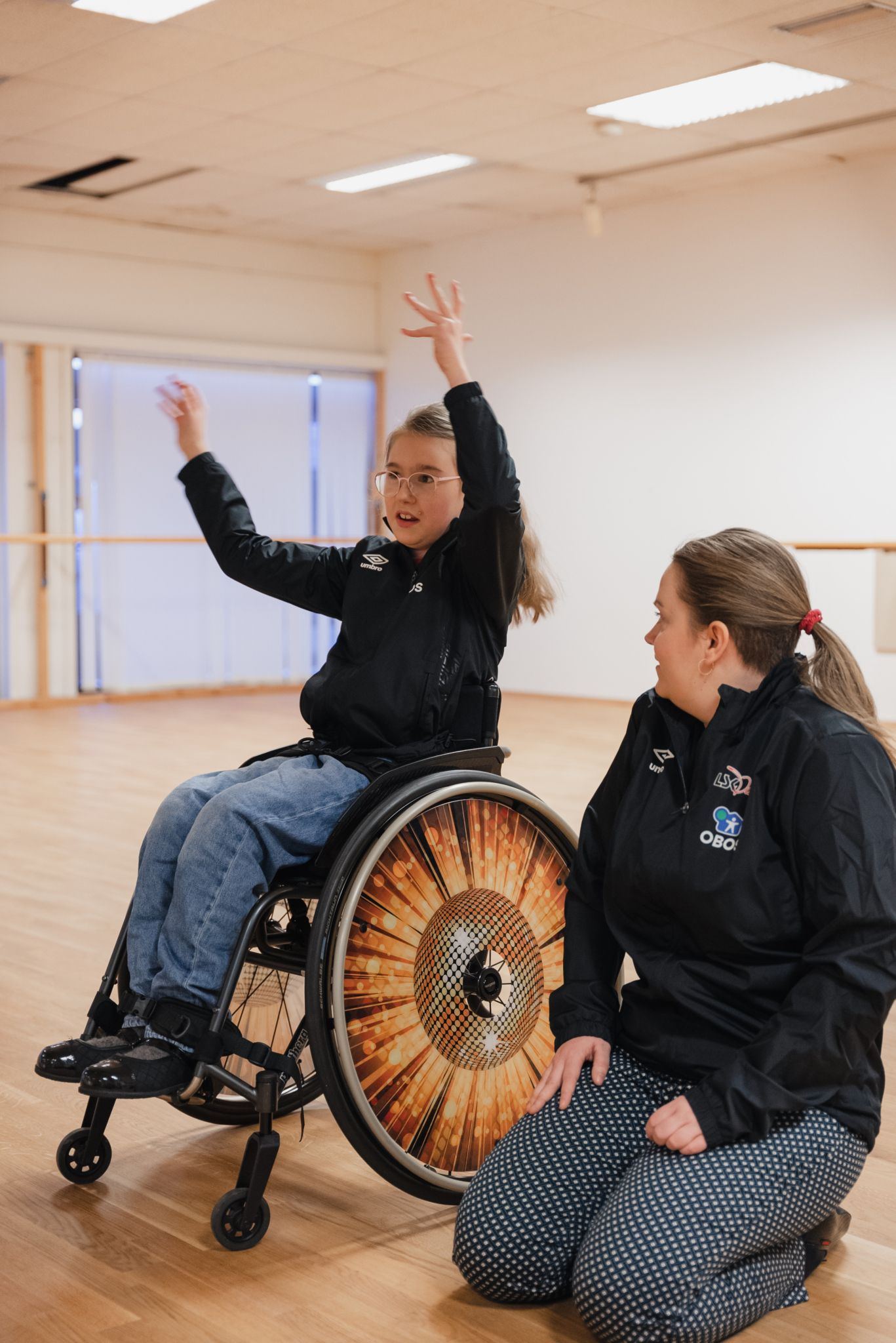 En jente i rullestol rekker armene i været. Til høyre for henne sitter en av instruktørene på knærne.