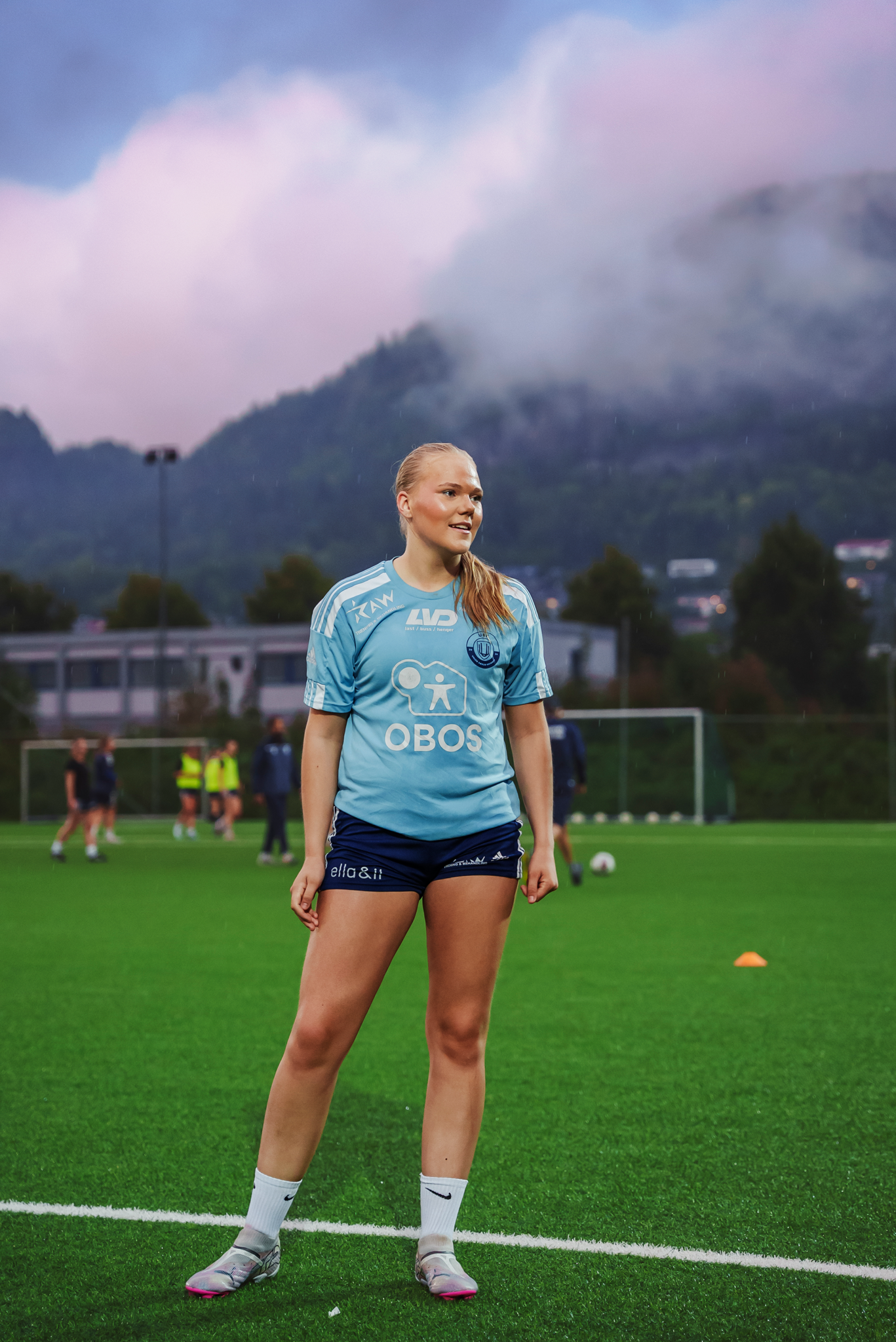 Othilie står og smiler ute på fotballbanen, med rosa skyer over henne.