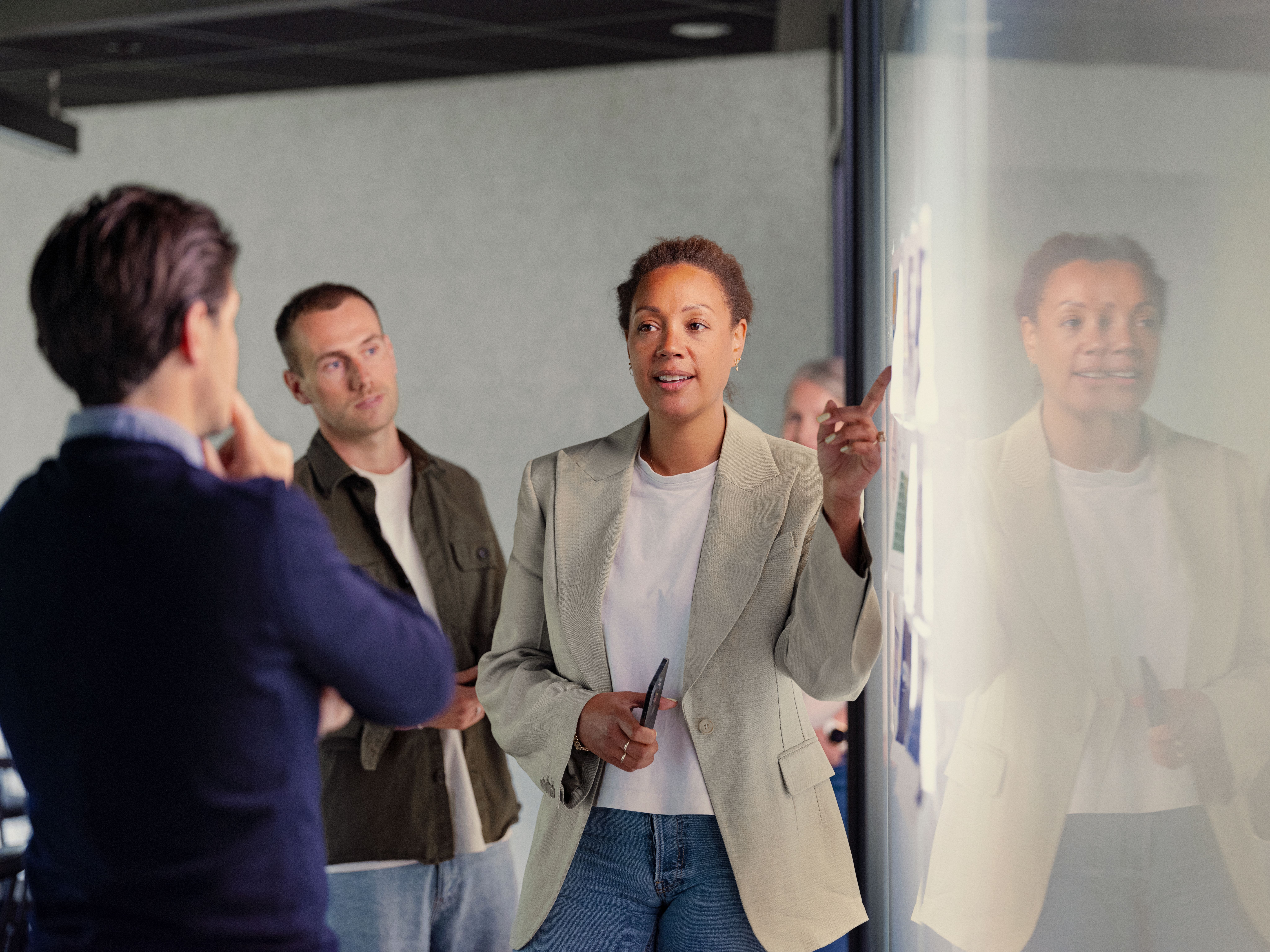 En kvinne i blazer presenterer selvsikkert for to kolleger og gestikulerer mot en glasstavle med lapper. Stemningen er fokusert og samarbeidsvillig.