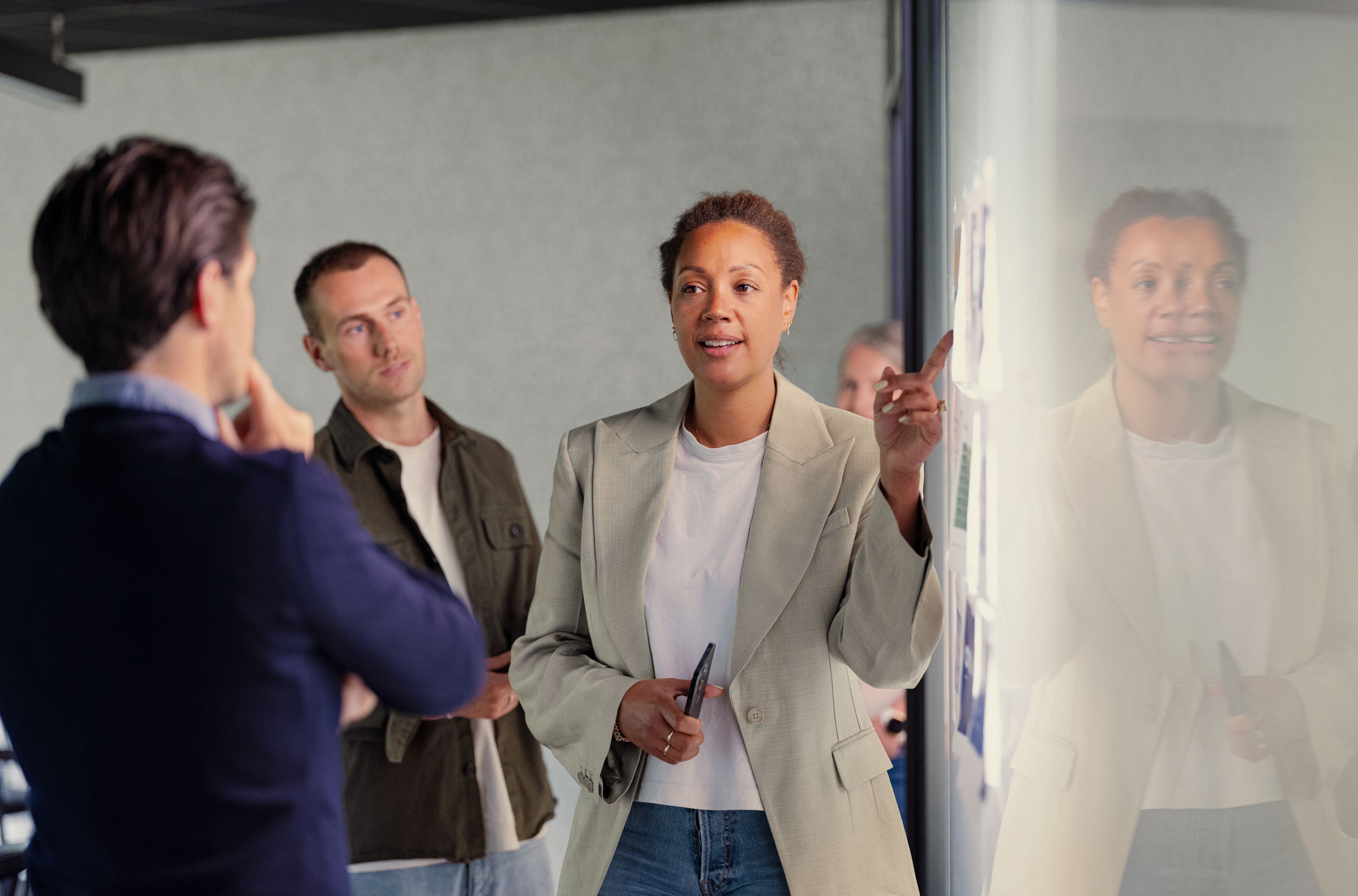 En kvinne i blazer presenterer selvsikkert for to kolleger og gestikulerer mot en glasstavle med lapper. Stemningen er fokusert og samarbeidsvillig.
