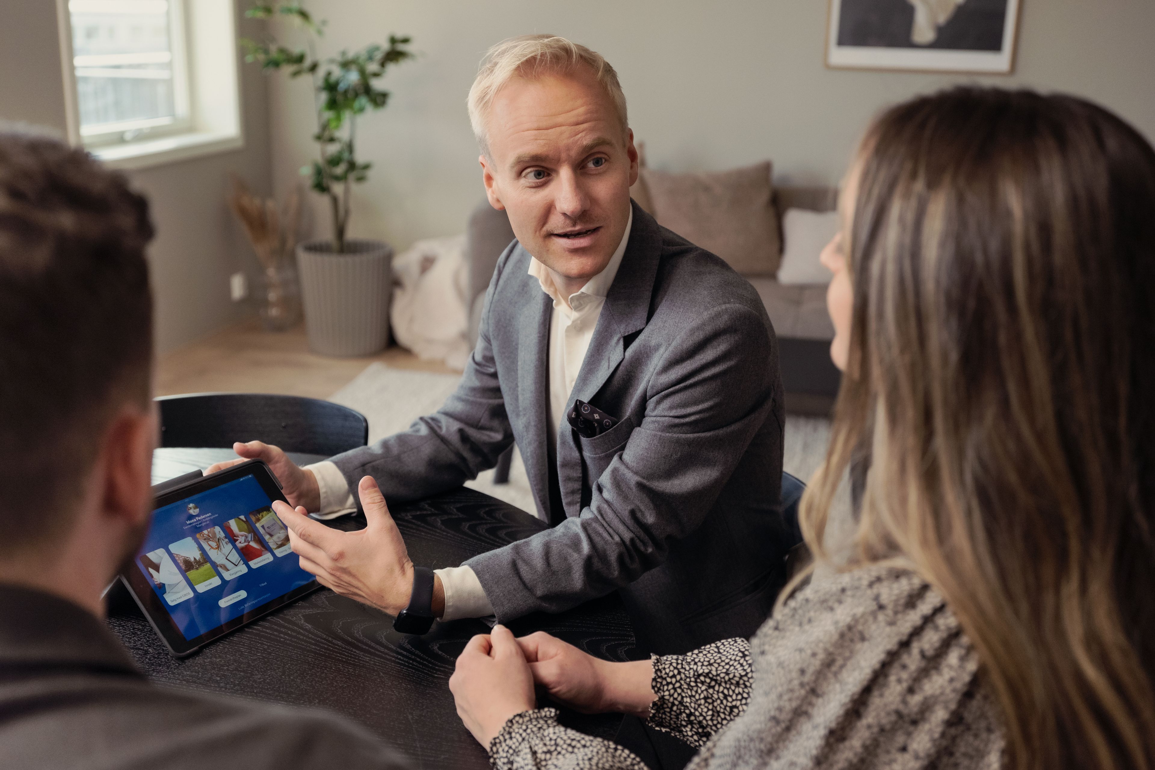 En mannlig eiendomsmegler viser fram informasjon på en iPad til en mann og en dame som sitter med ryggen til kameraet.