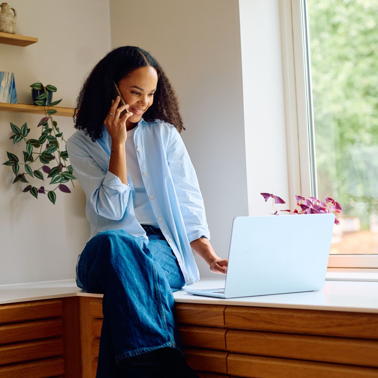 En smilende dame som sitter på benken og skriver på en Mac mens hun er i telefon.