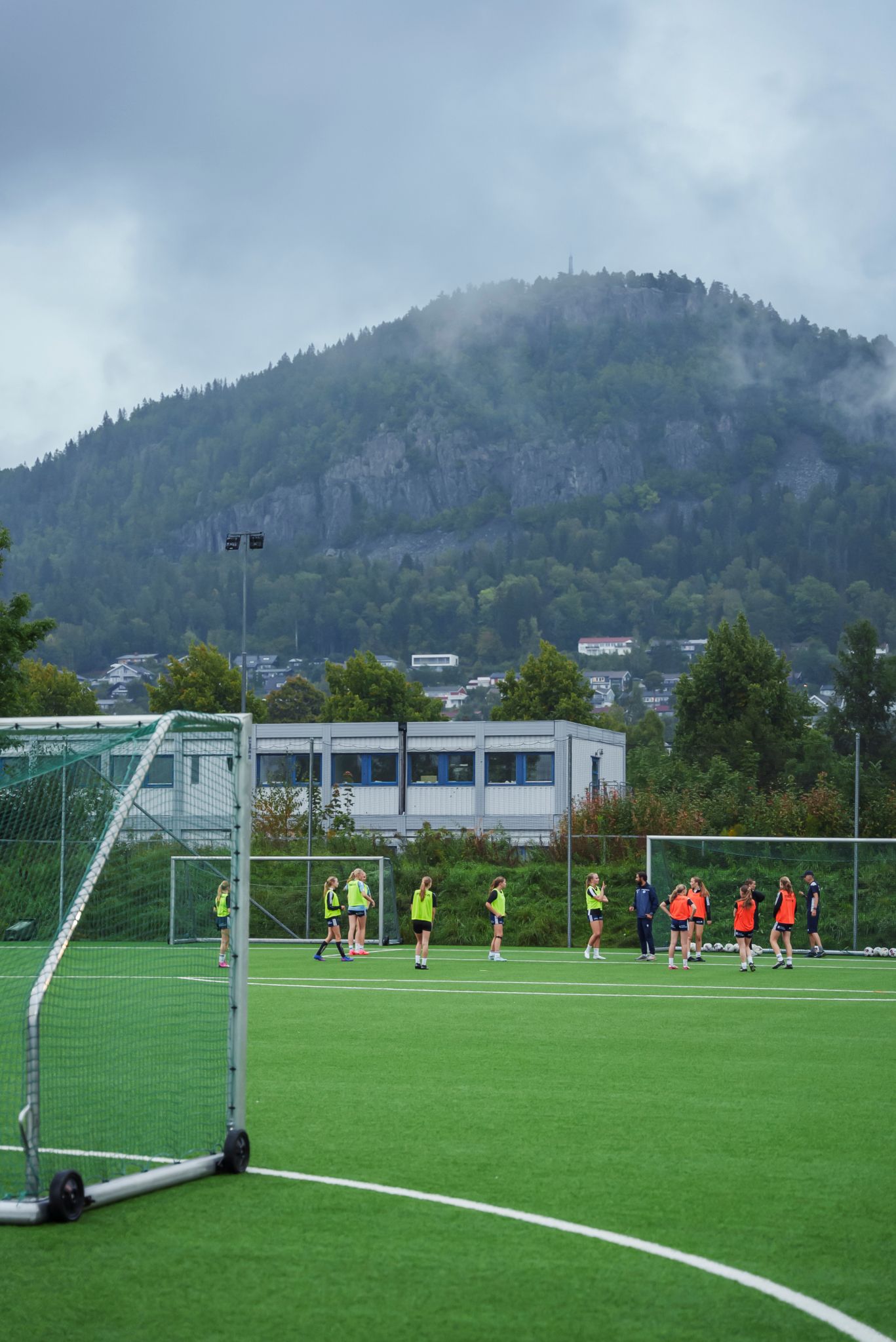 Bilde av en kunstgressbane mot et fjell i bakgrunnen. På andre siden av banen står jenter i vester og trener.