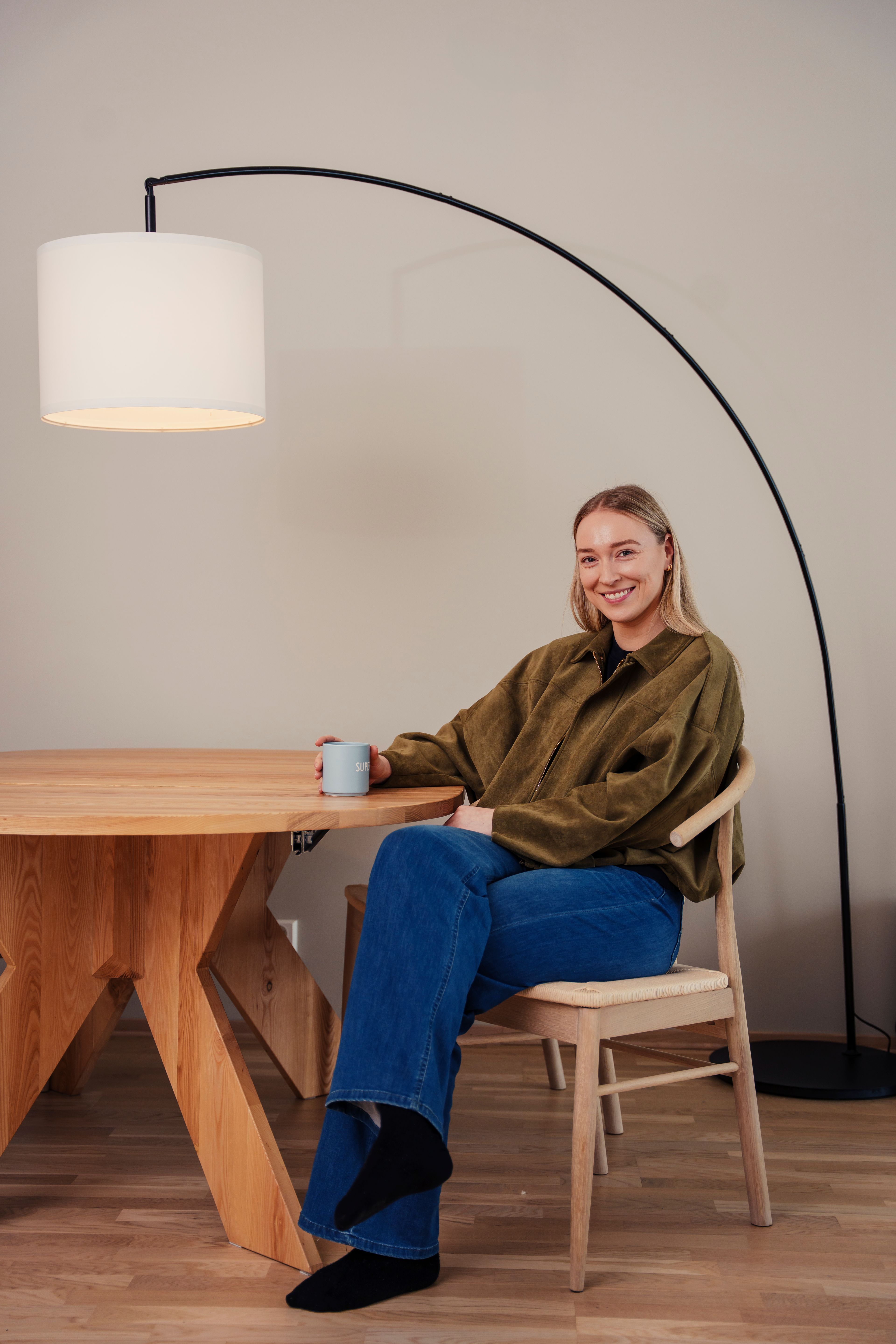 Eli Marie Raasok sitter ved et spisebord i tre, under en stor gulvlampe, og smiler mot kamera.