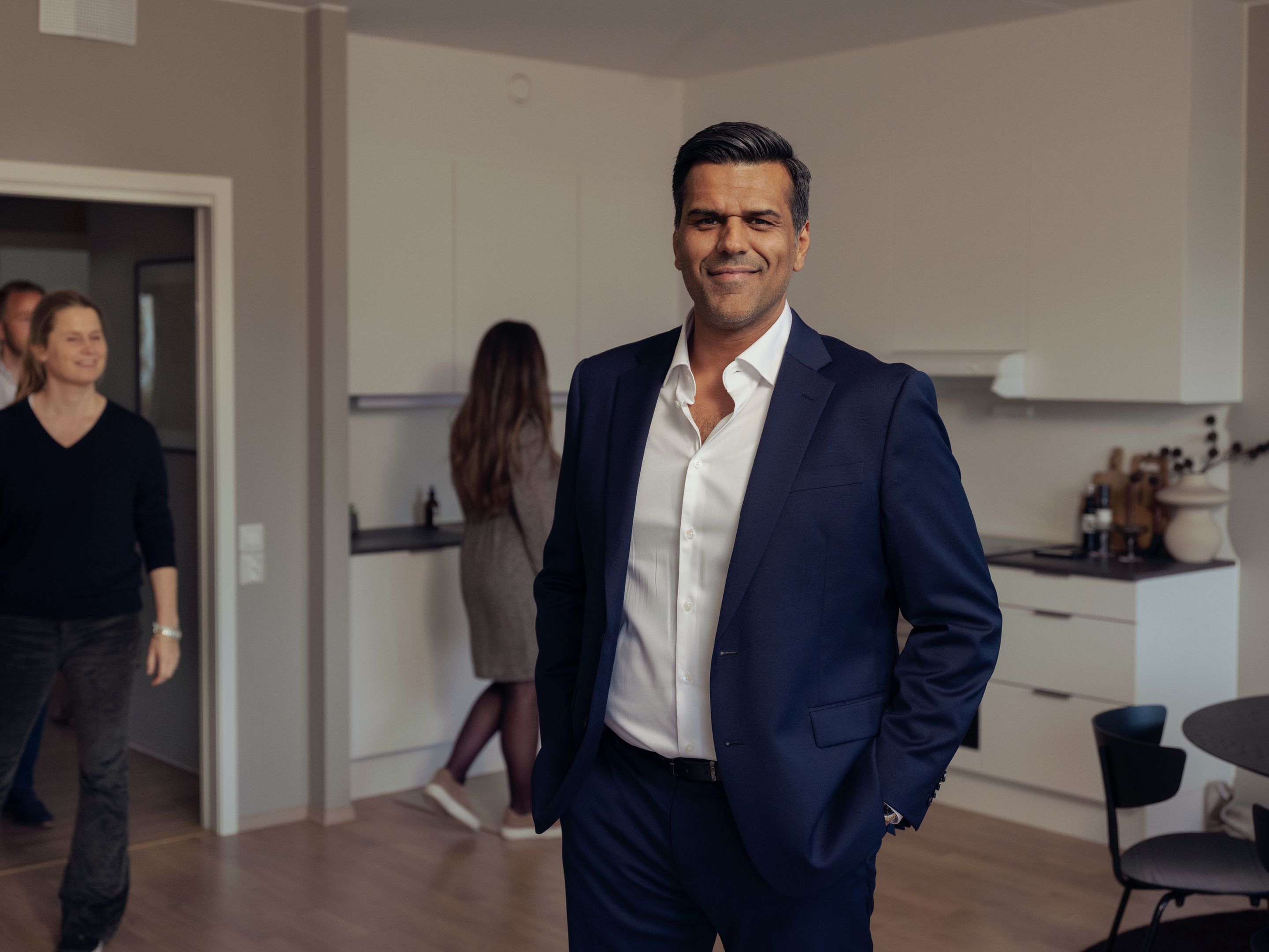 Naveed Iqbal, eiendomsmegler i OBOS Eiendomsmeglere, står i en leilighet med åpen stue- og kjøkkenløsning. Han smiler mot kameraet. I bakgrunnen er det tre personer på visning.
