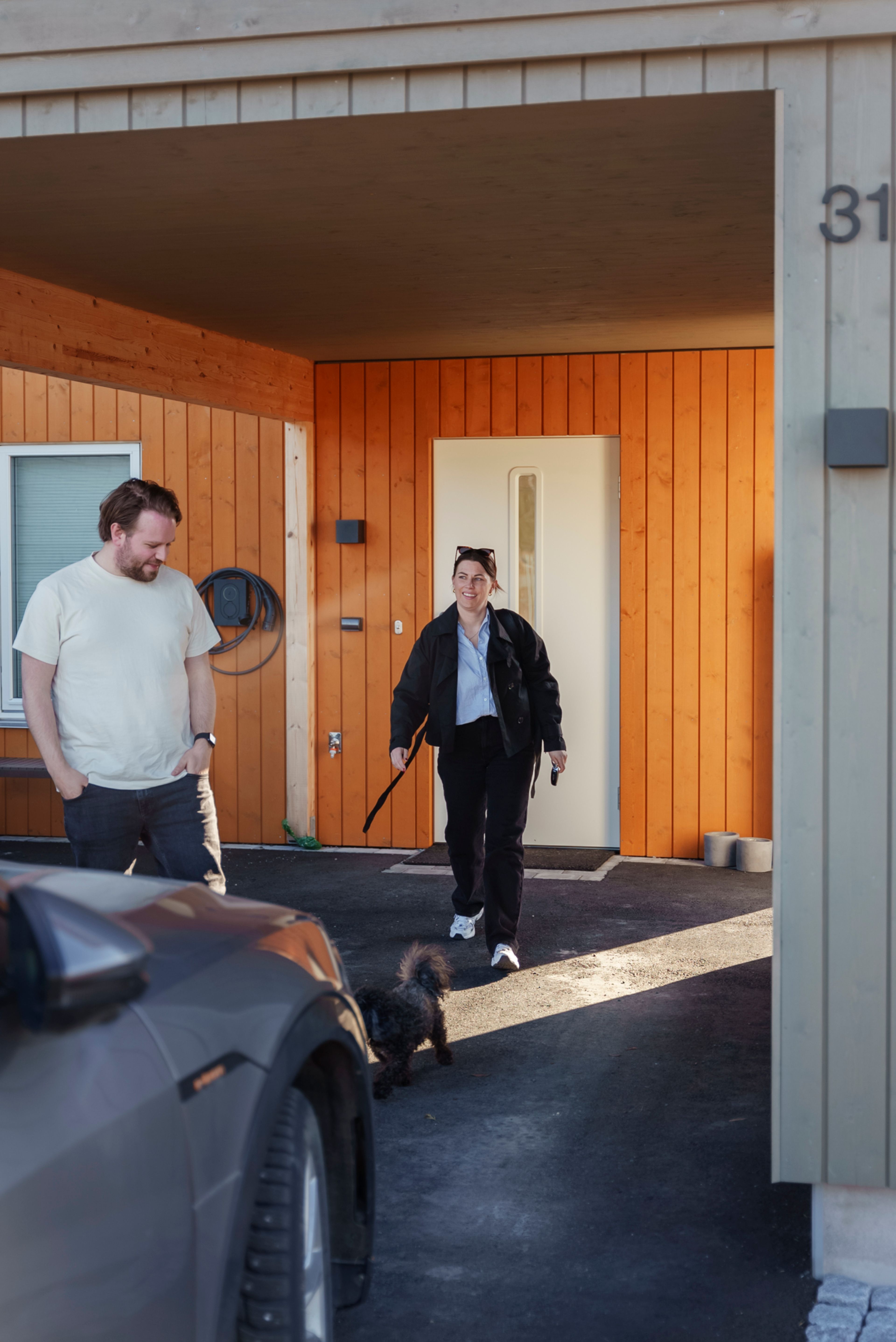 Stine og Martin på vei ut fra døren deres, med de oransje veggene på huset godt synlige. Bilen i garasjen skimtes foran dem