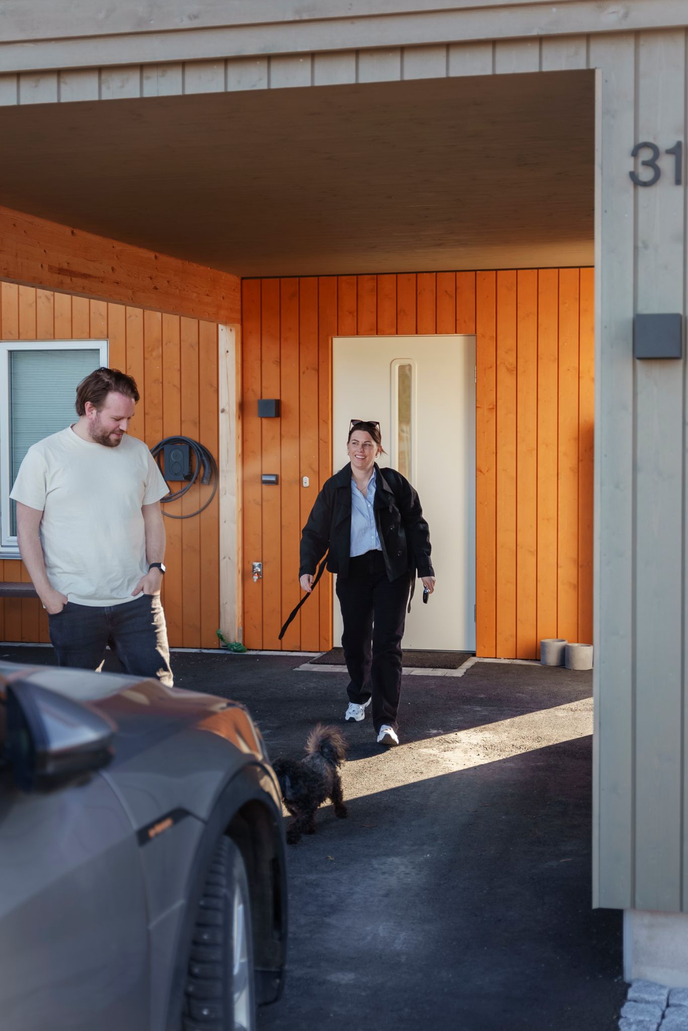 Stine og Martin på vei ut fra døren deres, med de oransje veggene på huset godt synlige. Bilen i garasjen skimtes foran dem