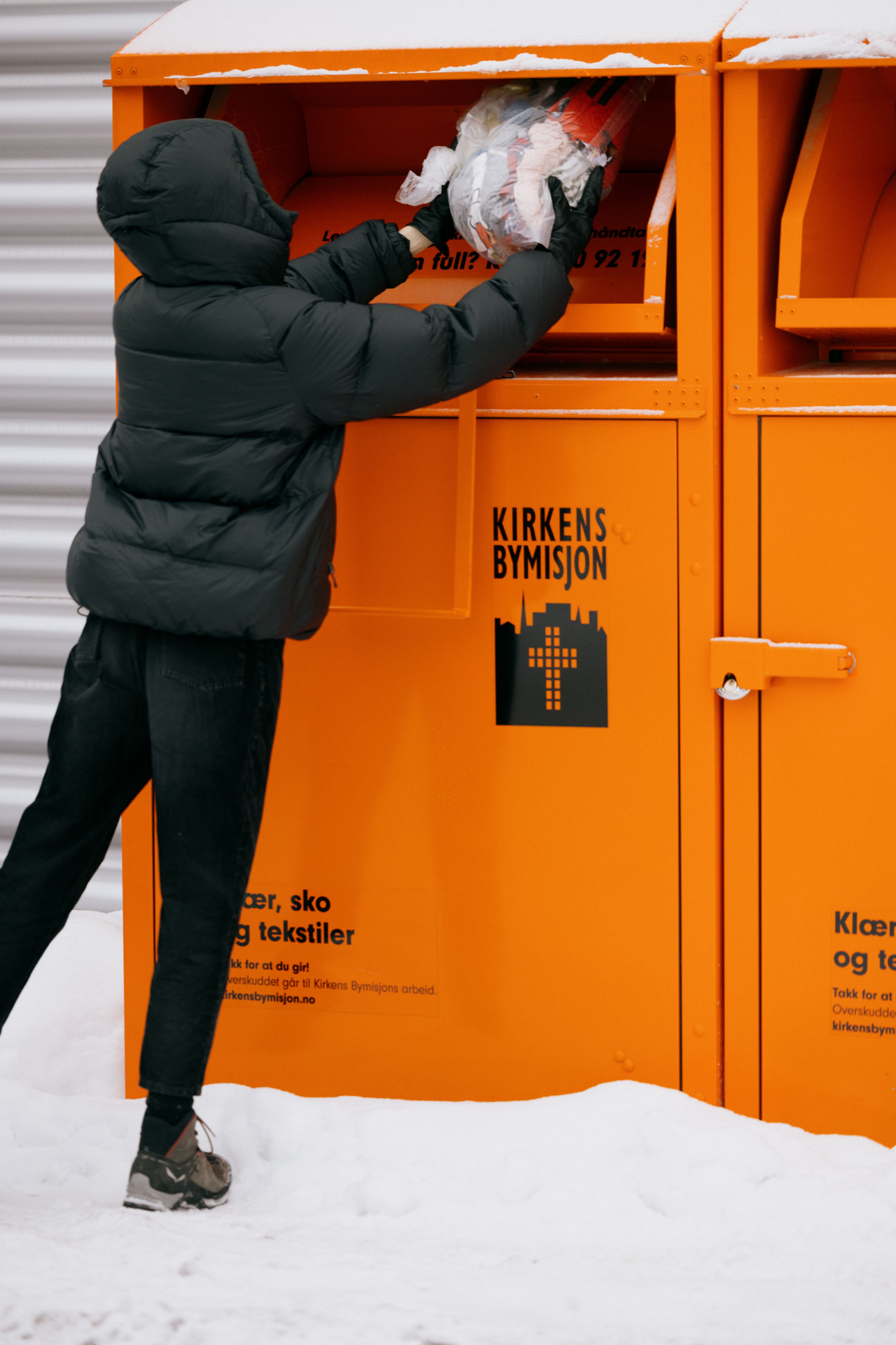 Foto av en person i sort dunjakke som kaster en poste med tøy i Kirkens Bymisjon sine oransje innsamlingsbokser.