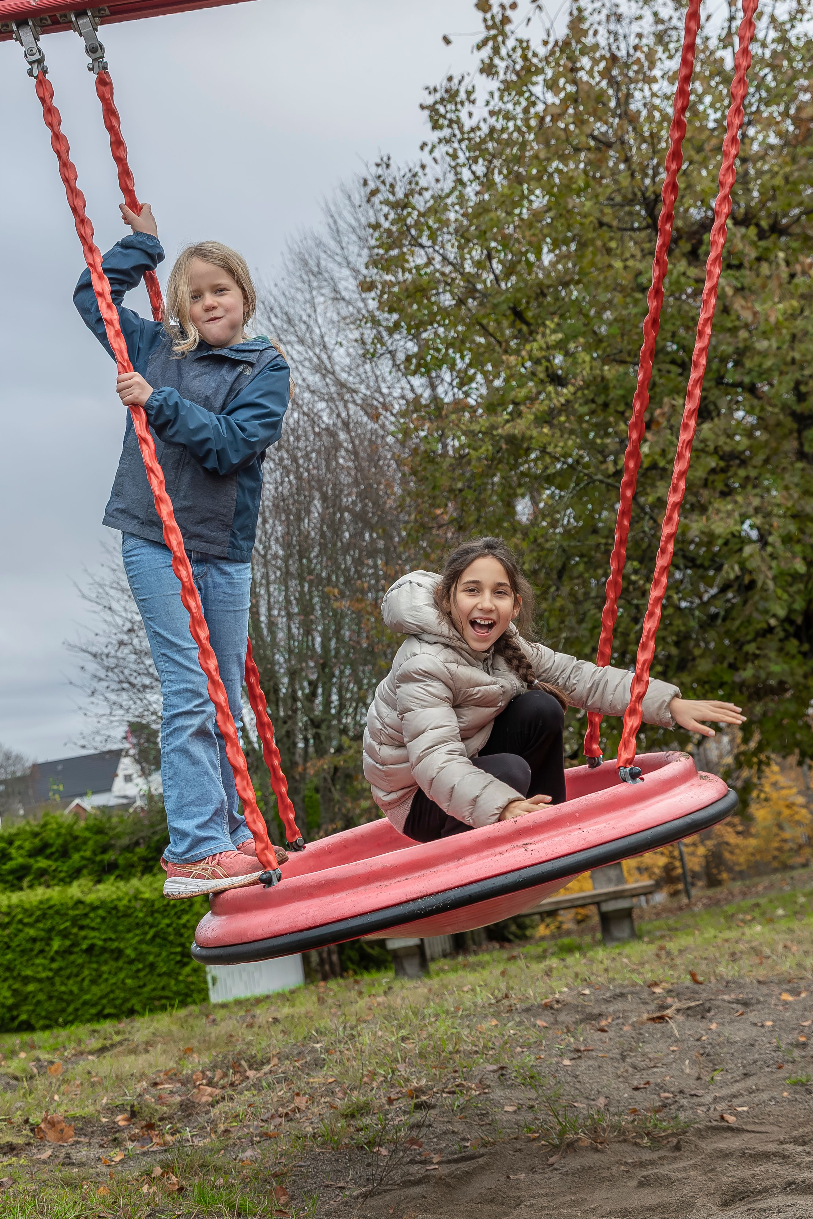 To barn leker på en stor rød huske i Stovner borettslag. Den ene står og holder i tauene mens den andre sitter og smiler.