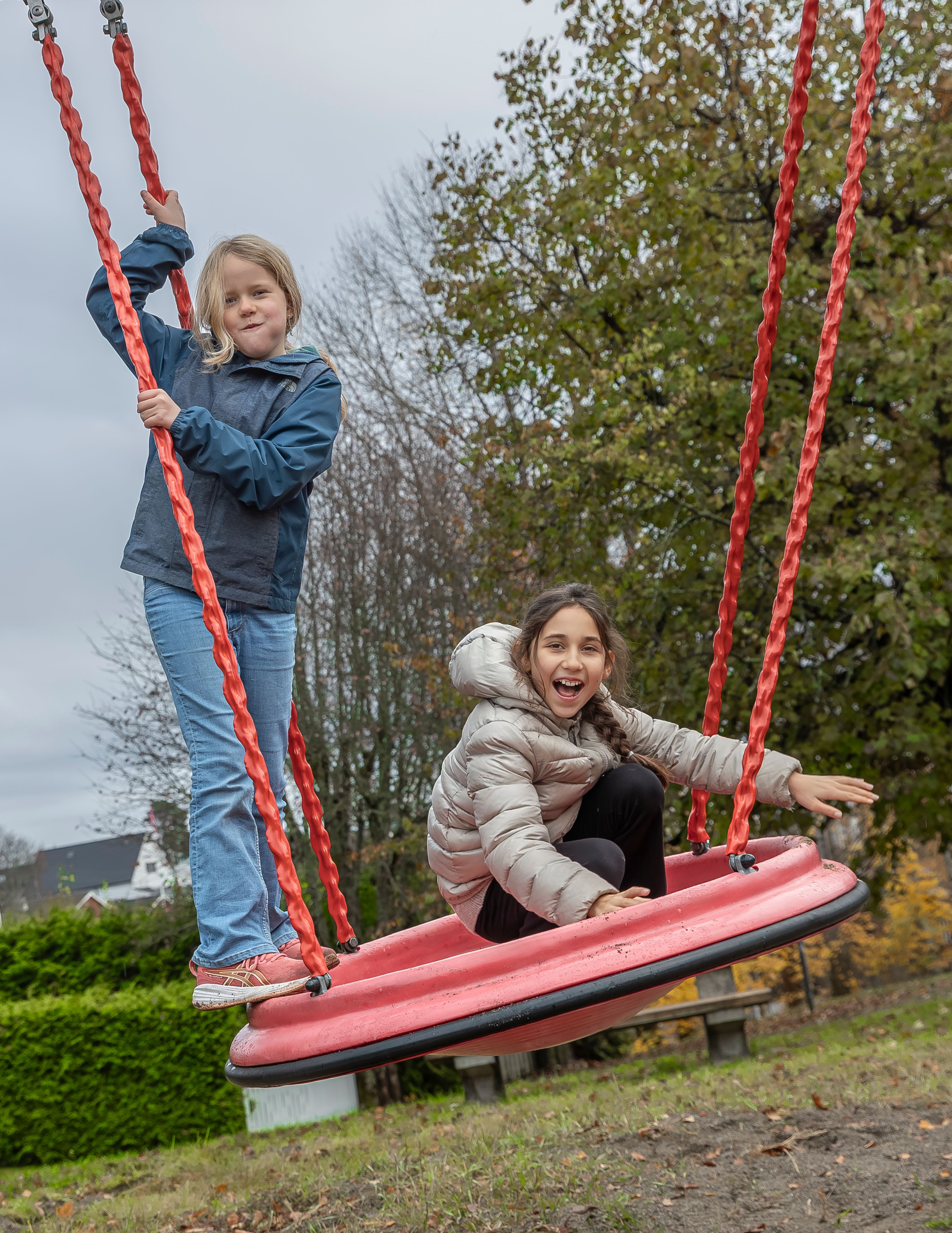 To barn leker på en stor rød huske i Stovner borettslag. Den ene står og holder i tauene mens den andre sitter og smiler.