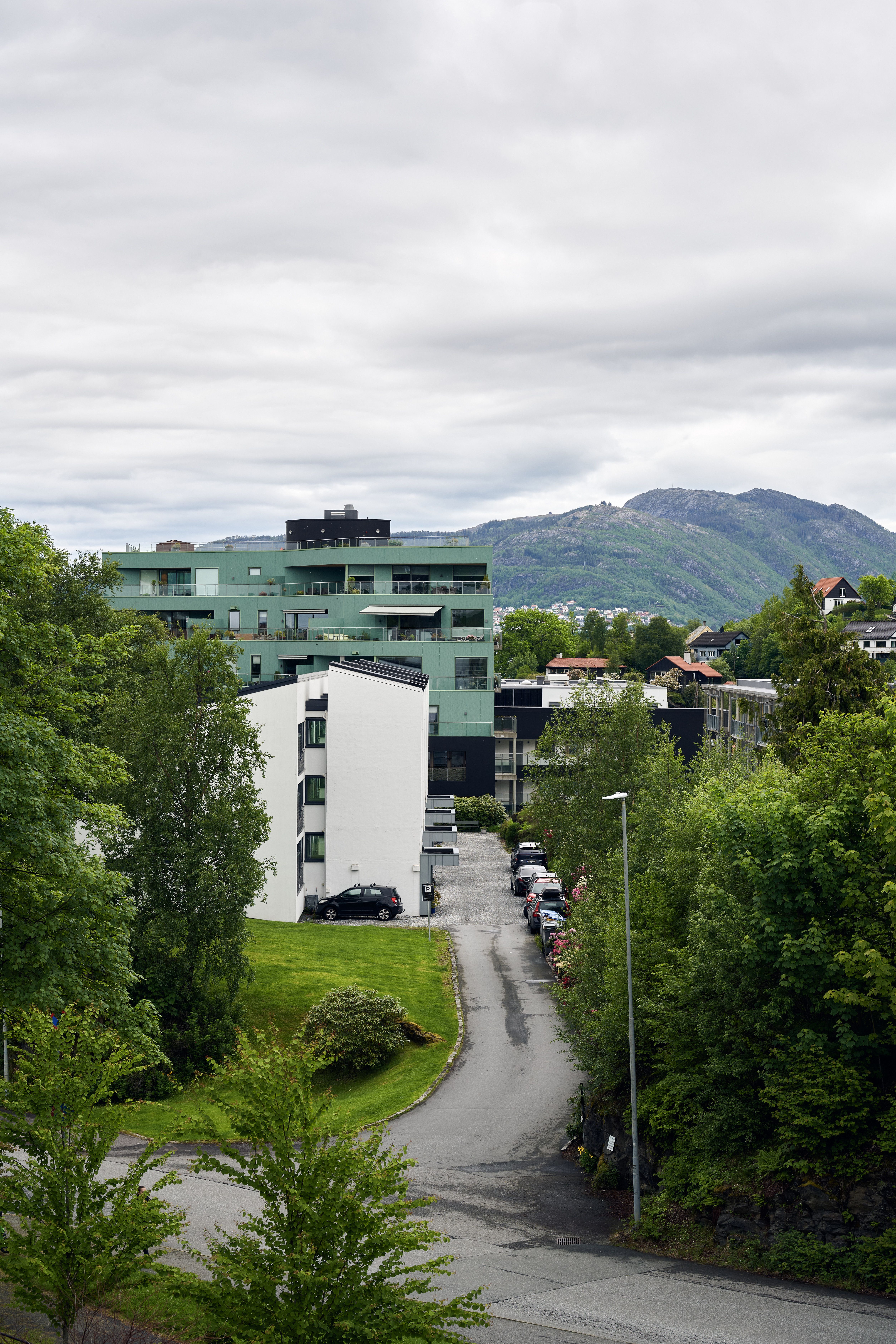 Grønn og hvit boligblokk på en skråning med en vei som snor seg ned mellom trær, og fjell i bakgrunnen.