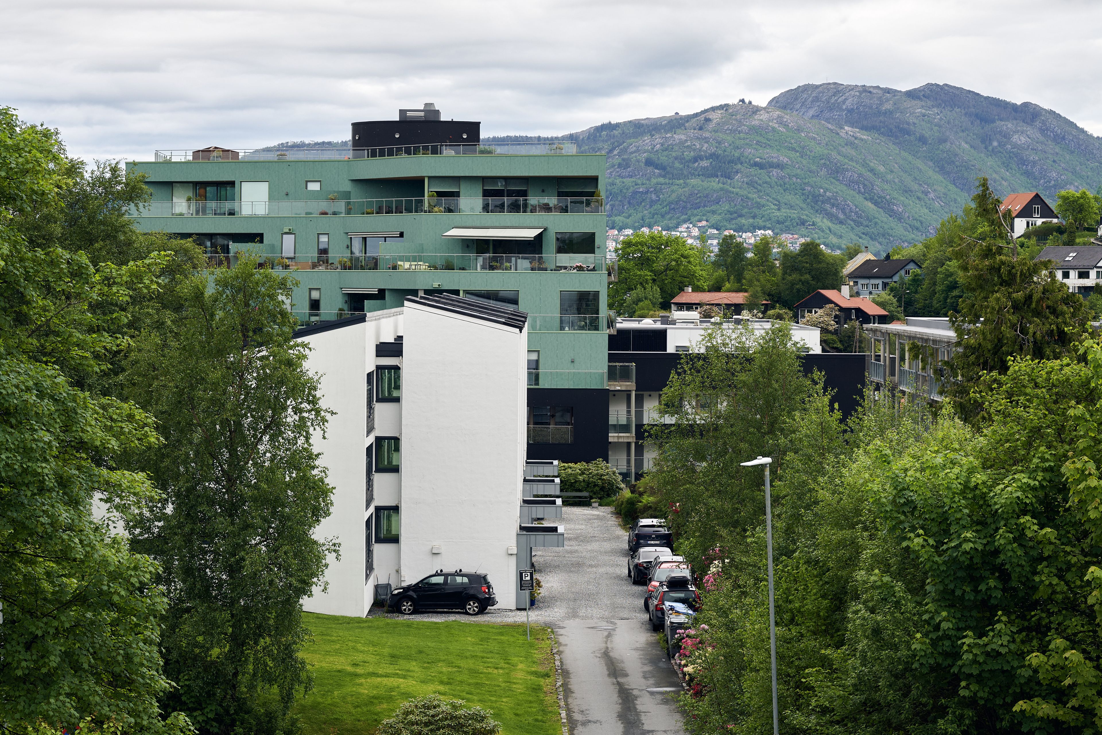 Grønn og hvit boligblokk på en skråning med en vei som snor seg ned mellom trær, og fjell i bakgrunnen.