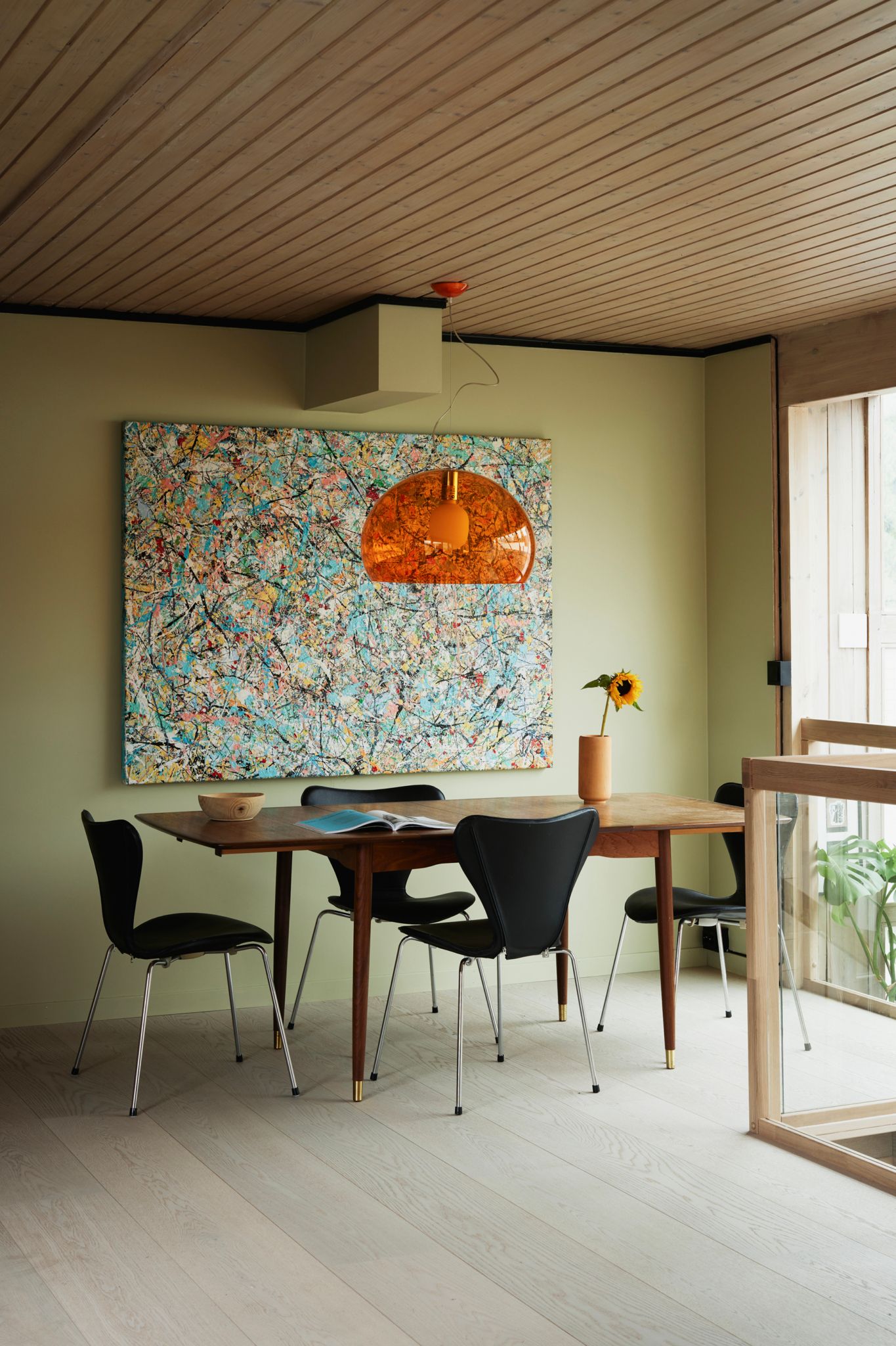 En spisekrok med et trebord, svarte stoler og et stort, fargerikt kunstverk på veggen. En oransje pendellampe henger over bordet, og en solsikke i en vase.