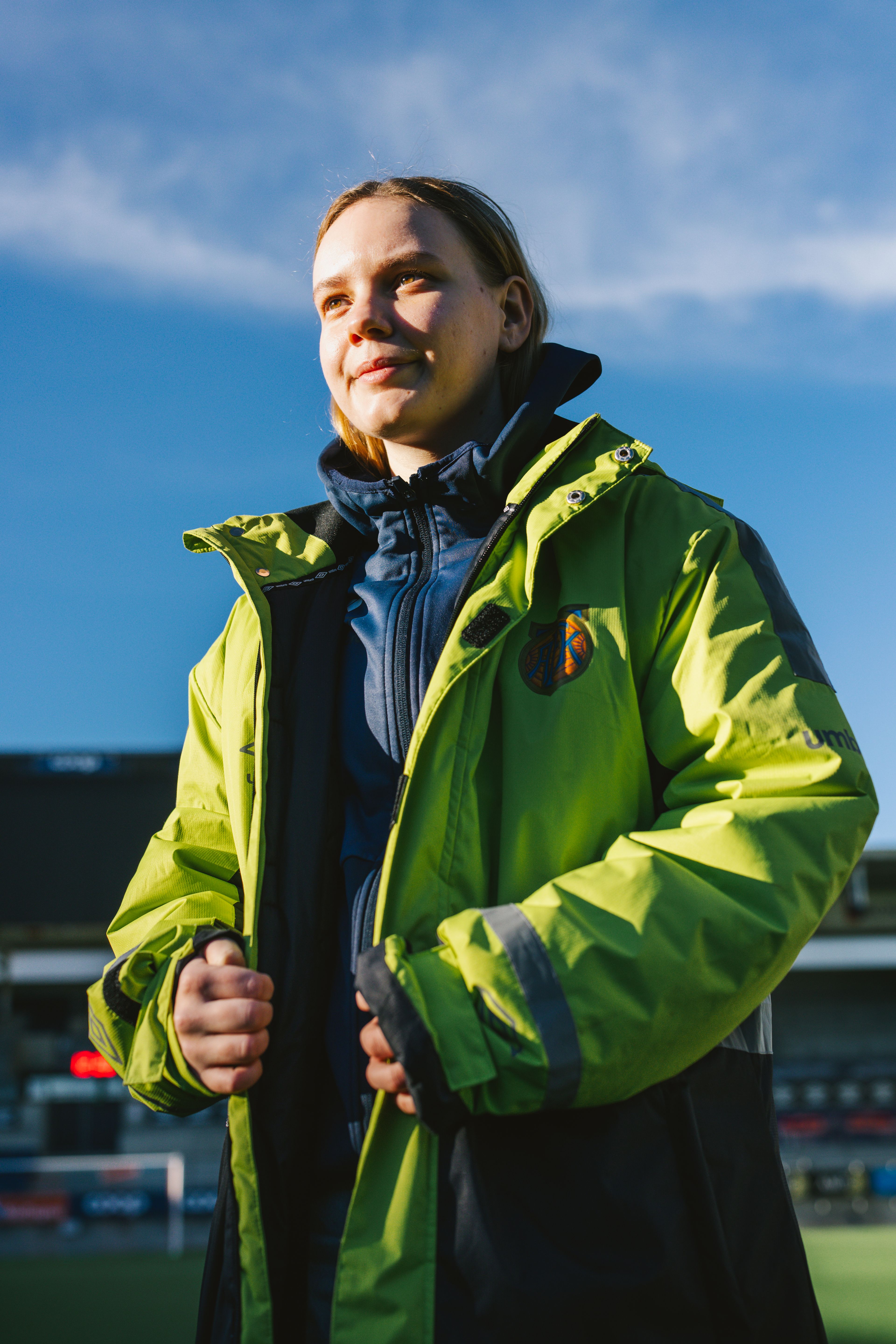 Nært portrett av Kristina i neongul kampvert-jakke utendørs på Color Line Stadion, med blå himmel bak.
