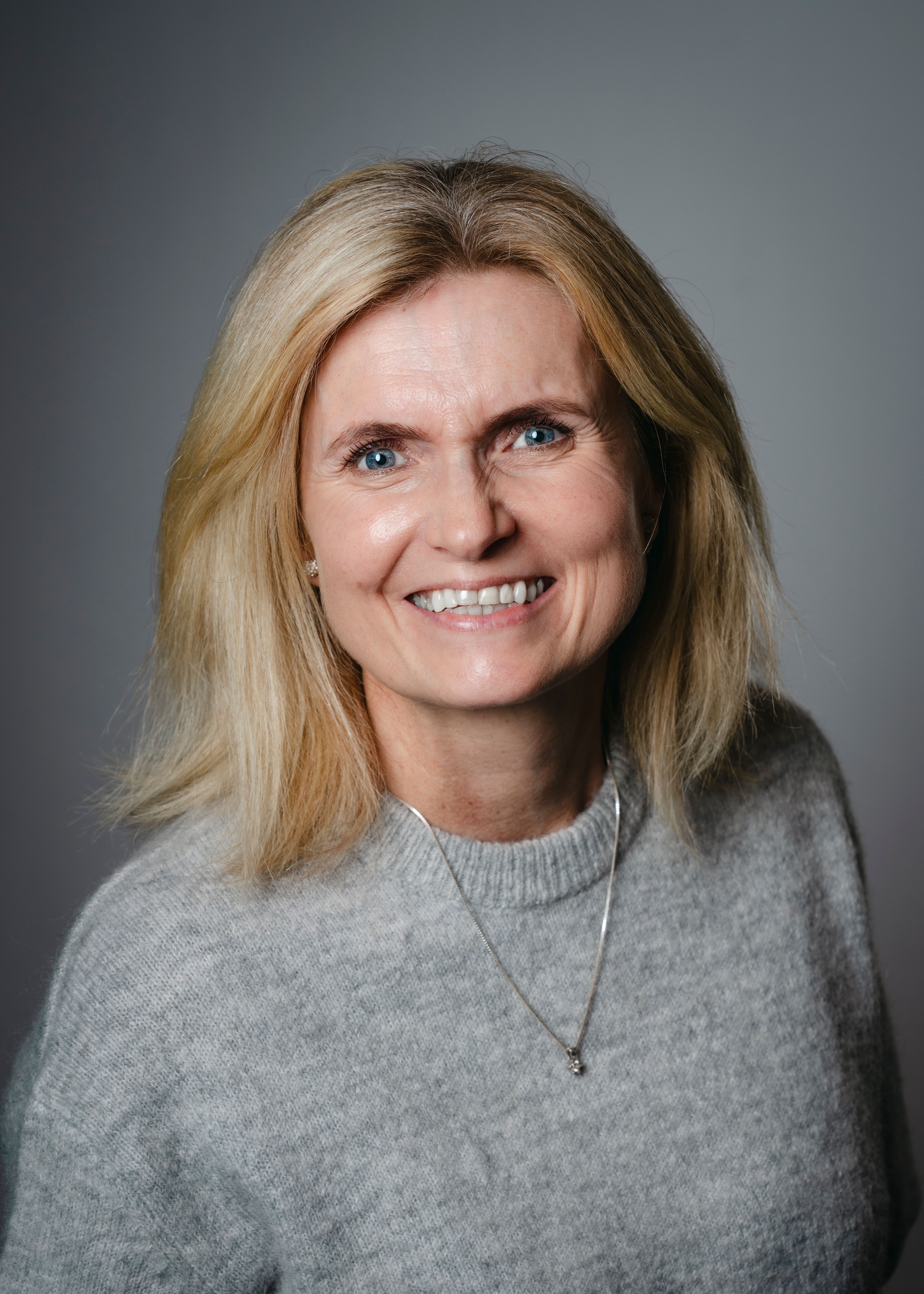 Smilende blond kvinne i grå genser fotografert mot en nøytral grå studiobakgrunn.