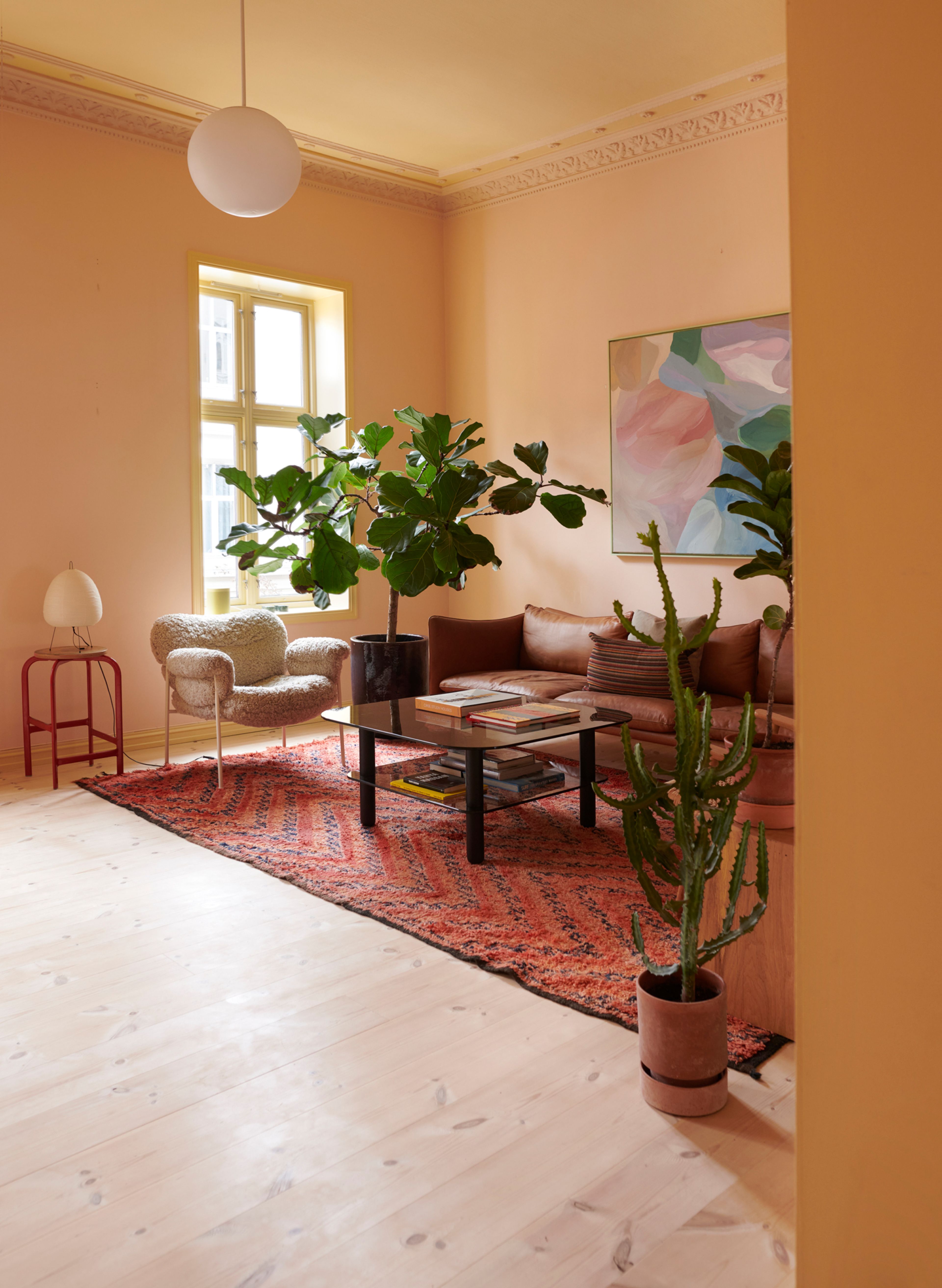Stue malt i en dus, rosa farge. En kaktus står i forgrunnen og bak ser vi ulike stue møbler, belysning, planter og et vindu.