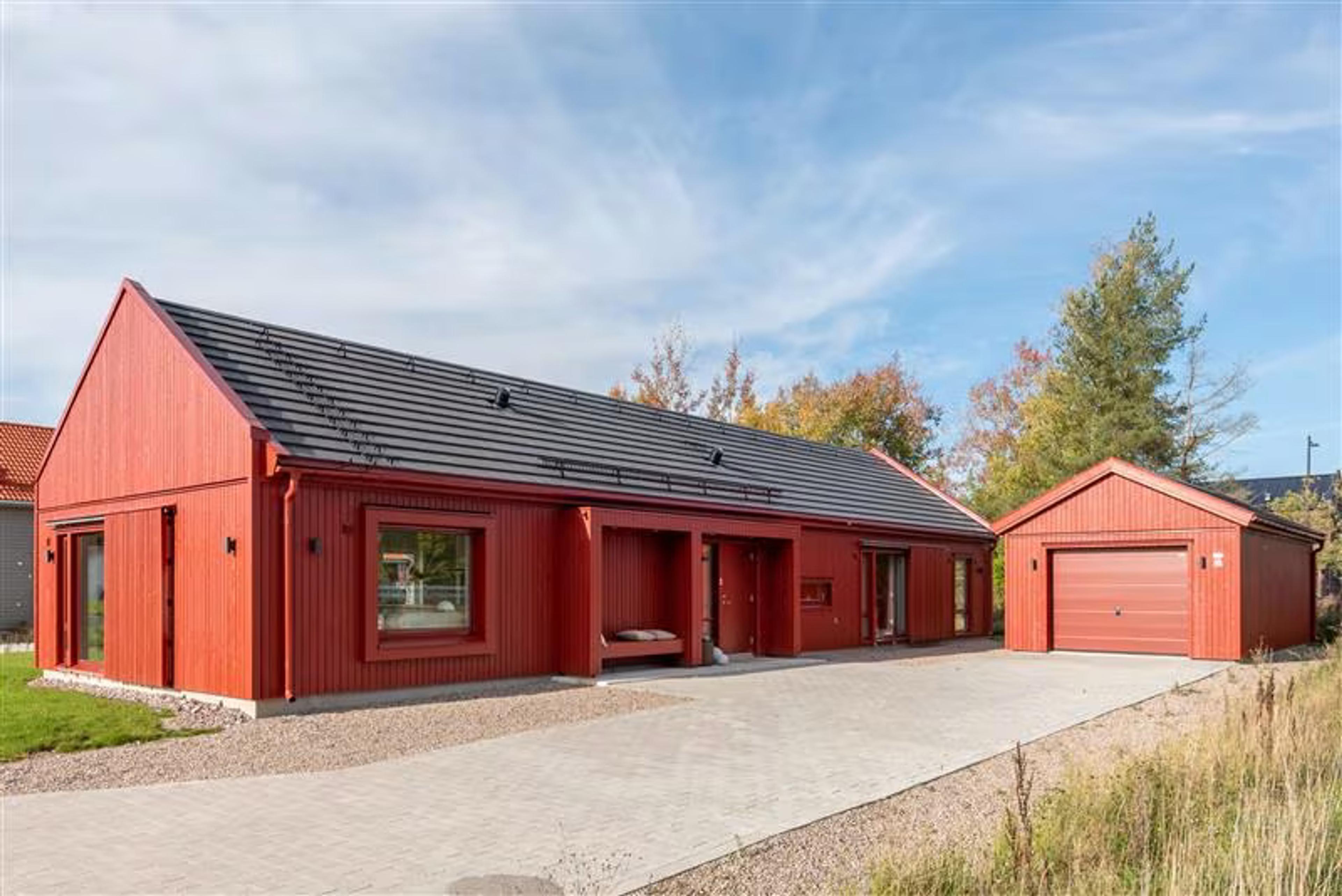 Et moderne rødt trehus med svart tak står ved siden av en separat garasje under en blå himmel. Høsttrær omgir eiendommen og skaper en fredelig atmosfære.