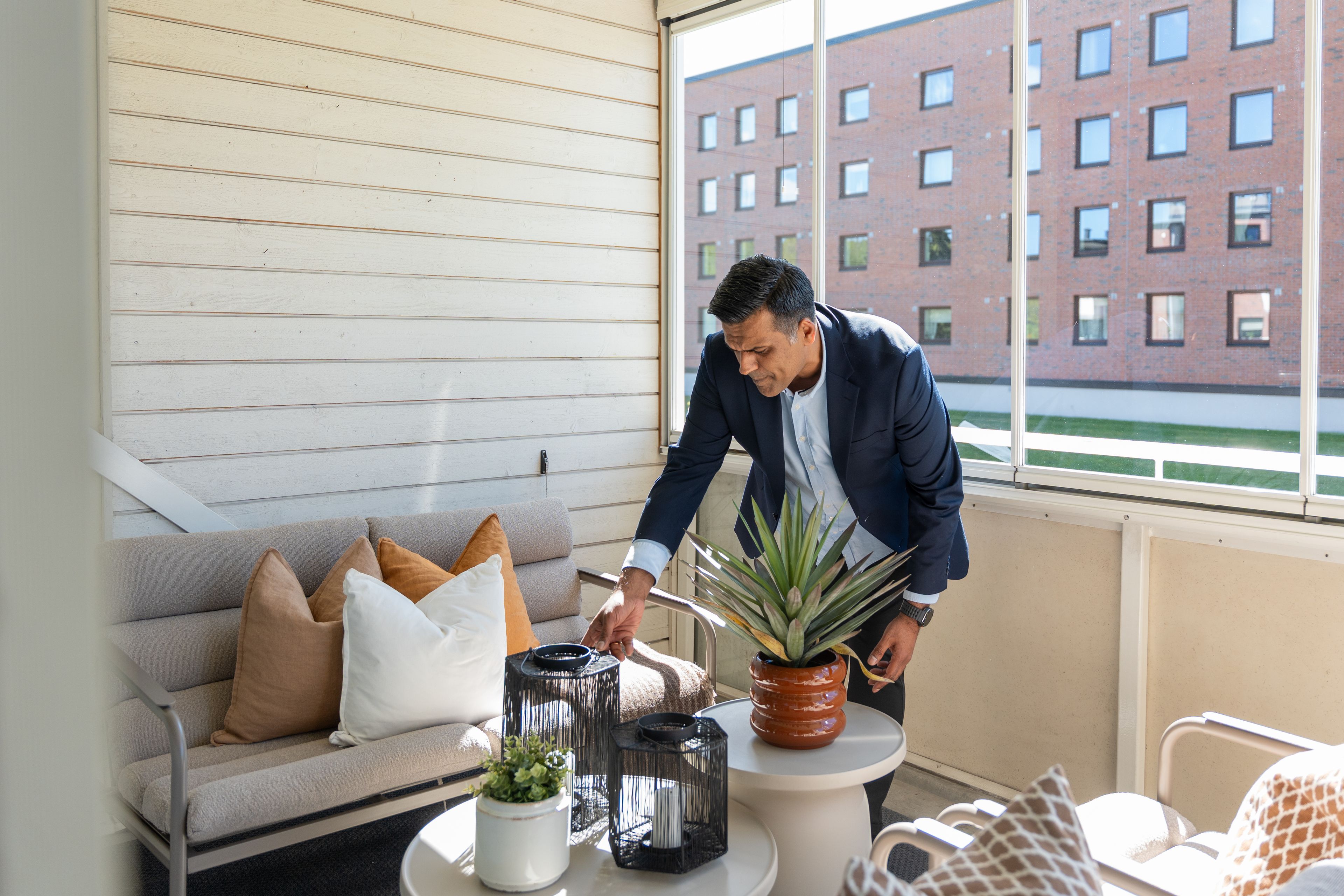 En mann i mørk blazer lener seg over et lite bord på en innglasset veranda og justerer en dekorasjon. På bordet står lykter og en agaveplante i en terrakottakrukke. I bakgrunnen ses en teglsteinsbygning gjennom vinduet.
