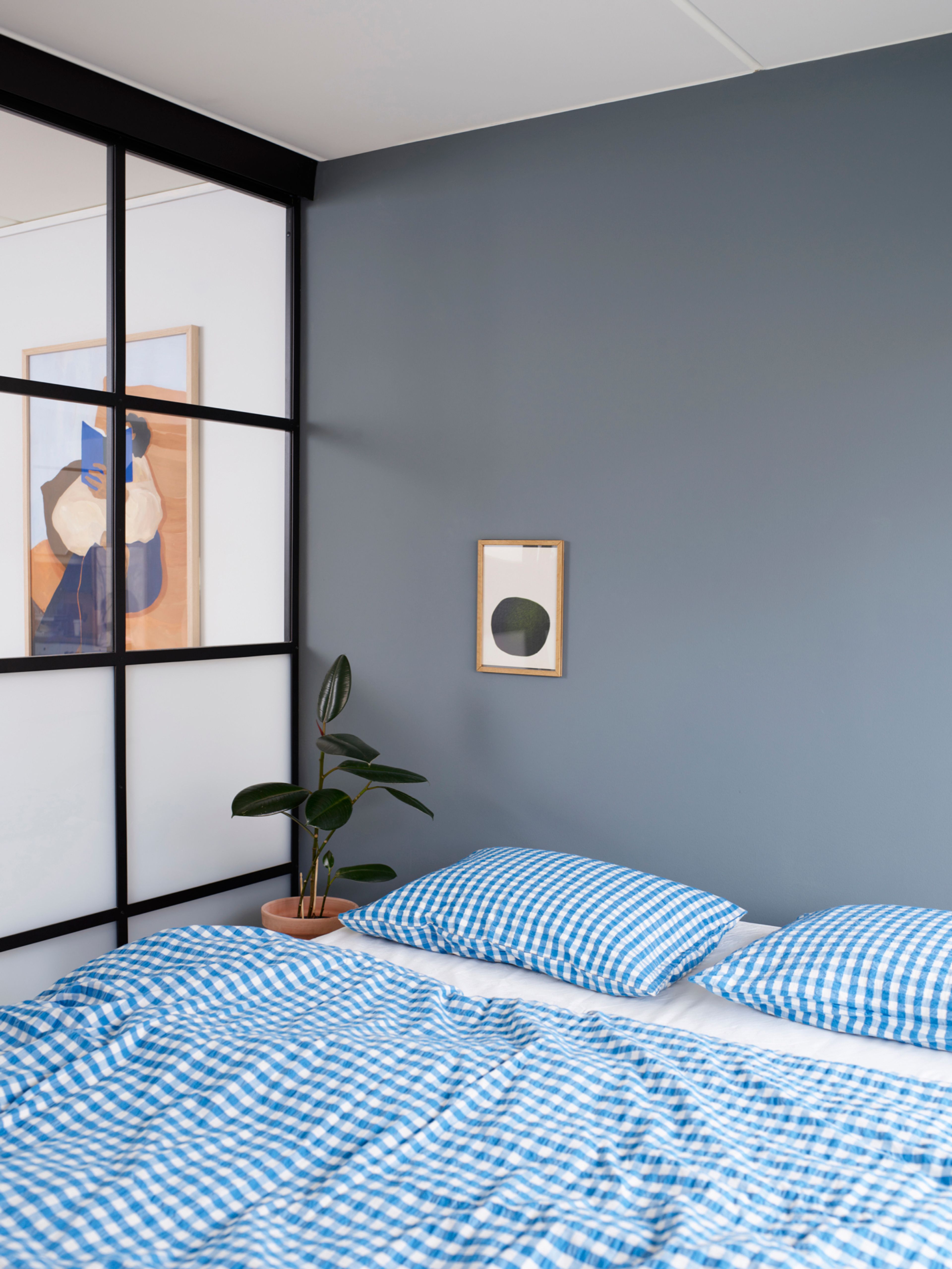 Blått soverom skilles fra stua med en industriell glassvegg, med seng med blårutete sengetrekk.