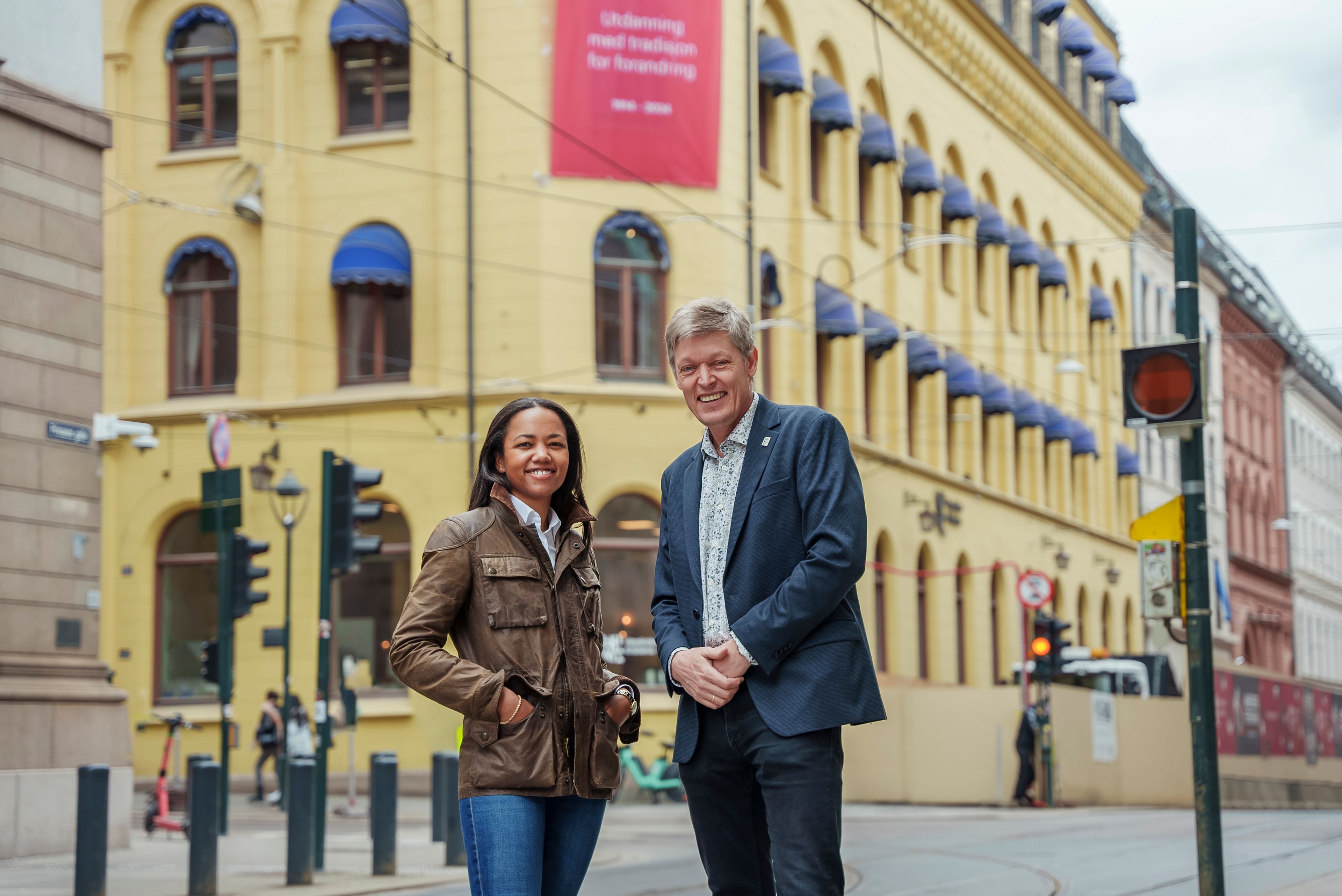 En kvinne og en mann står foran bygningen til Høyskolen Kristiania. De smiler. Bygget er gult.