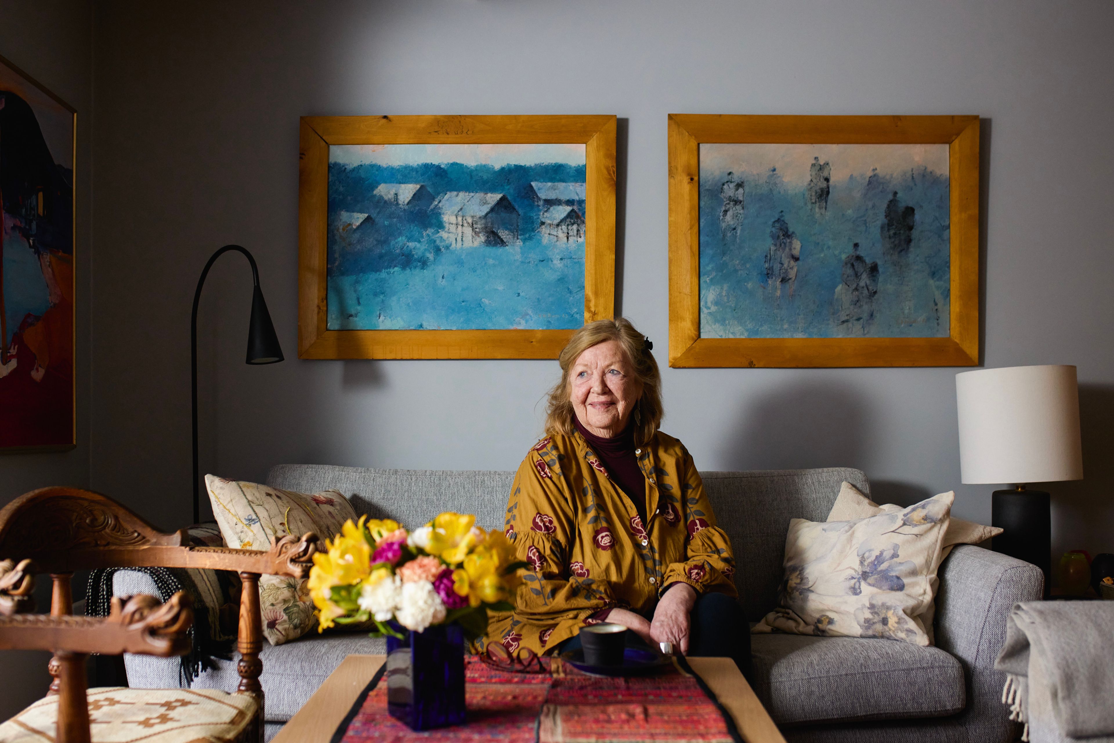 En kvinne sitter i sofaen sin hjemme. Hun titter ut og smiler. Det er to kunstverk på veggen bak henne og blomster i en vase på stuebordet.