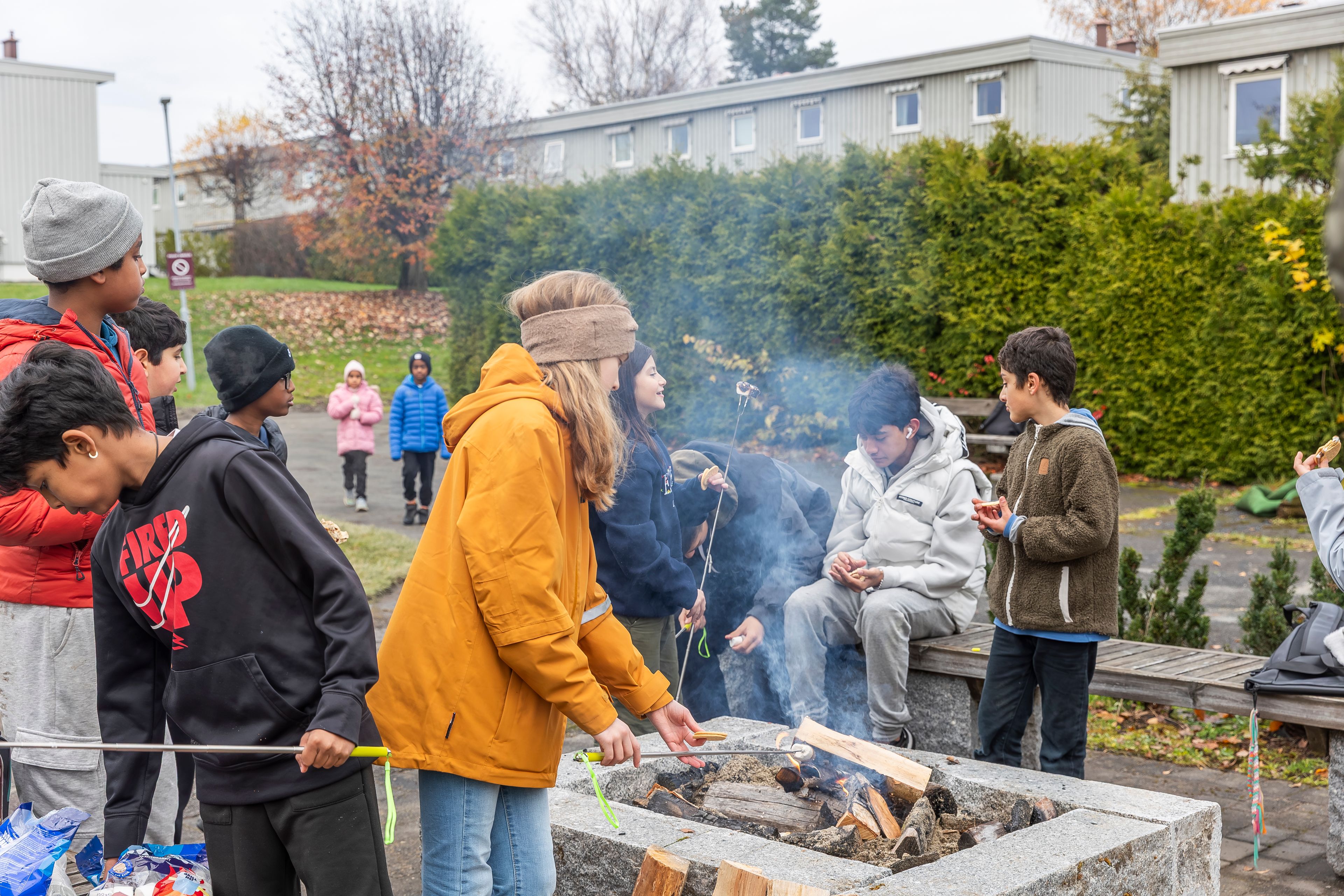 Barn og ungdom står rundt en bålpanne og griller marshmallows på uteområdet i Stovner borettslag.